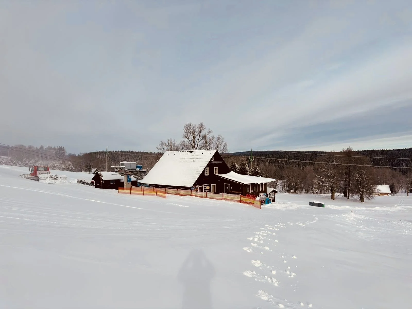 Snow-covered landscape with a black house and snow machinery, trees, and footprints in the snow under a cloudy sky.