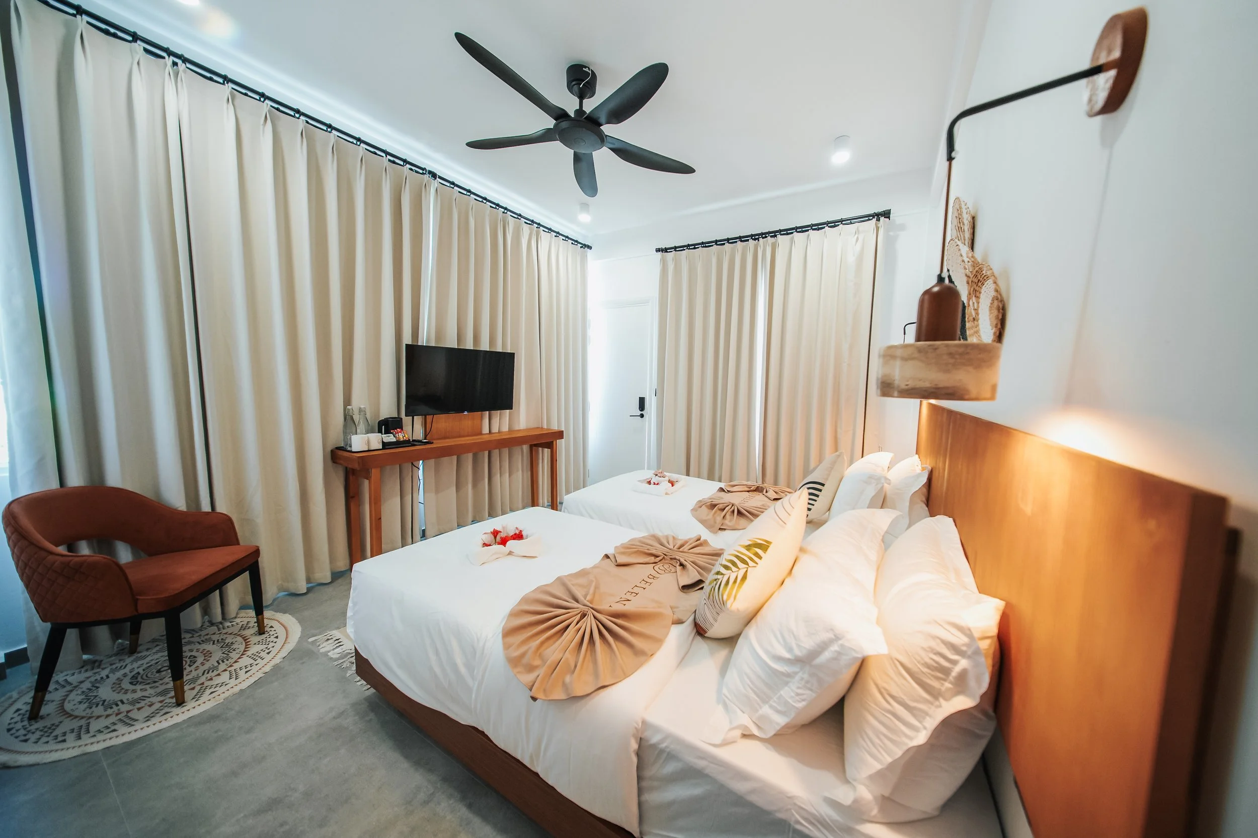 Hotel room with two beds, brown chair, wall-mounted TV, ceiling fan, and curtains. Bedspreads artfully arranged. Modern decor.