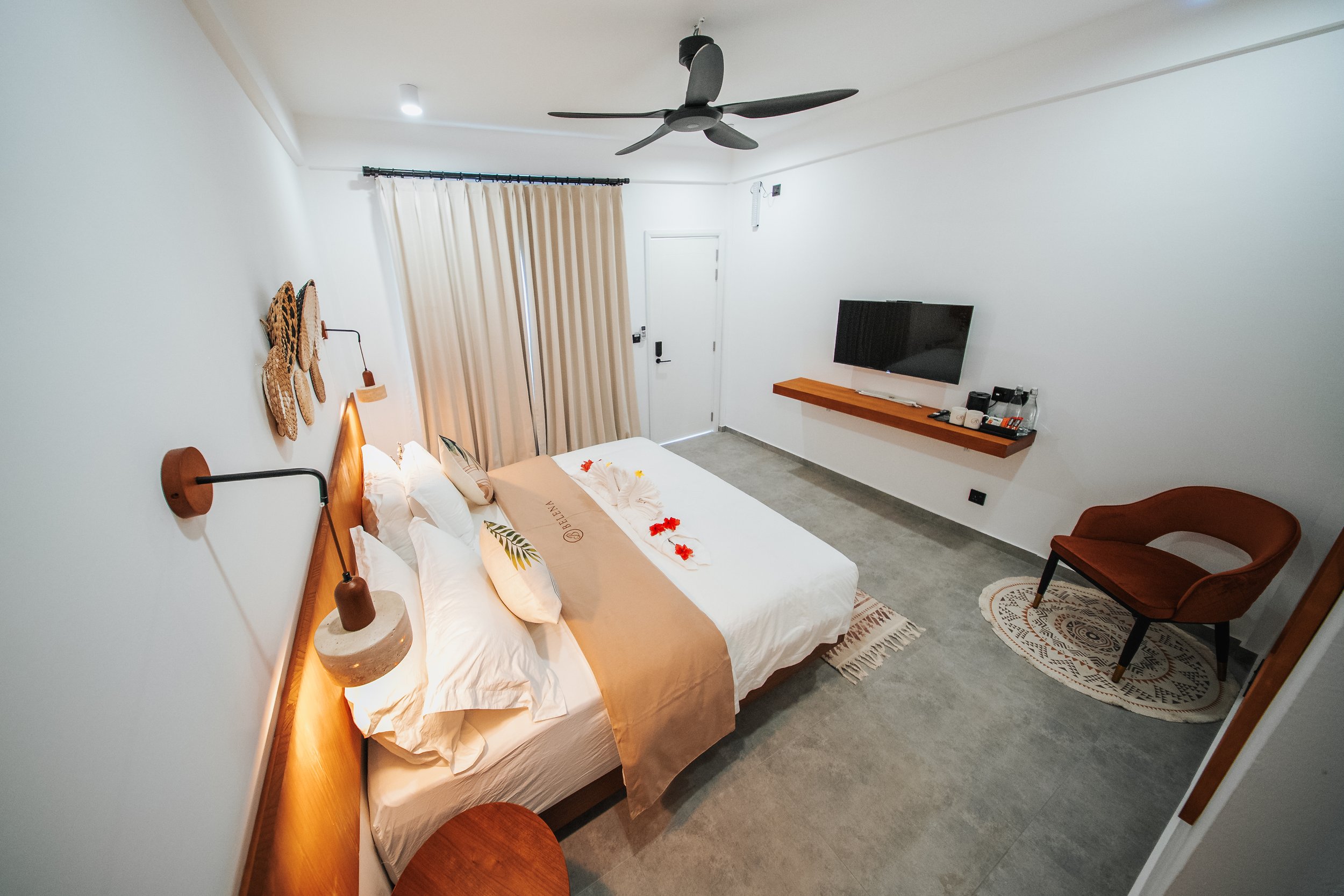 Modern bedroom with double bed, decorative pillows, and bedside lamps. A wall-mounted TV is above a floating shelf with drinks and snacks. A ceiling fan is above. White walls and floor with a brown accent chair on a round rug. Beige curtains cover th