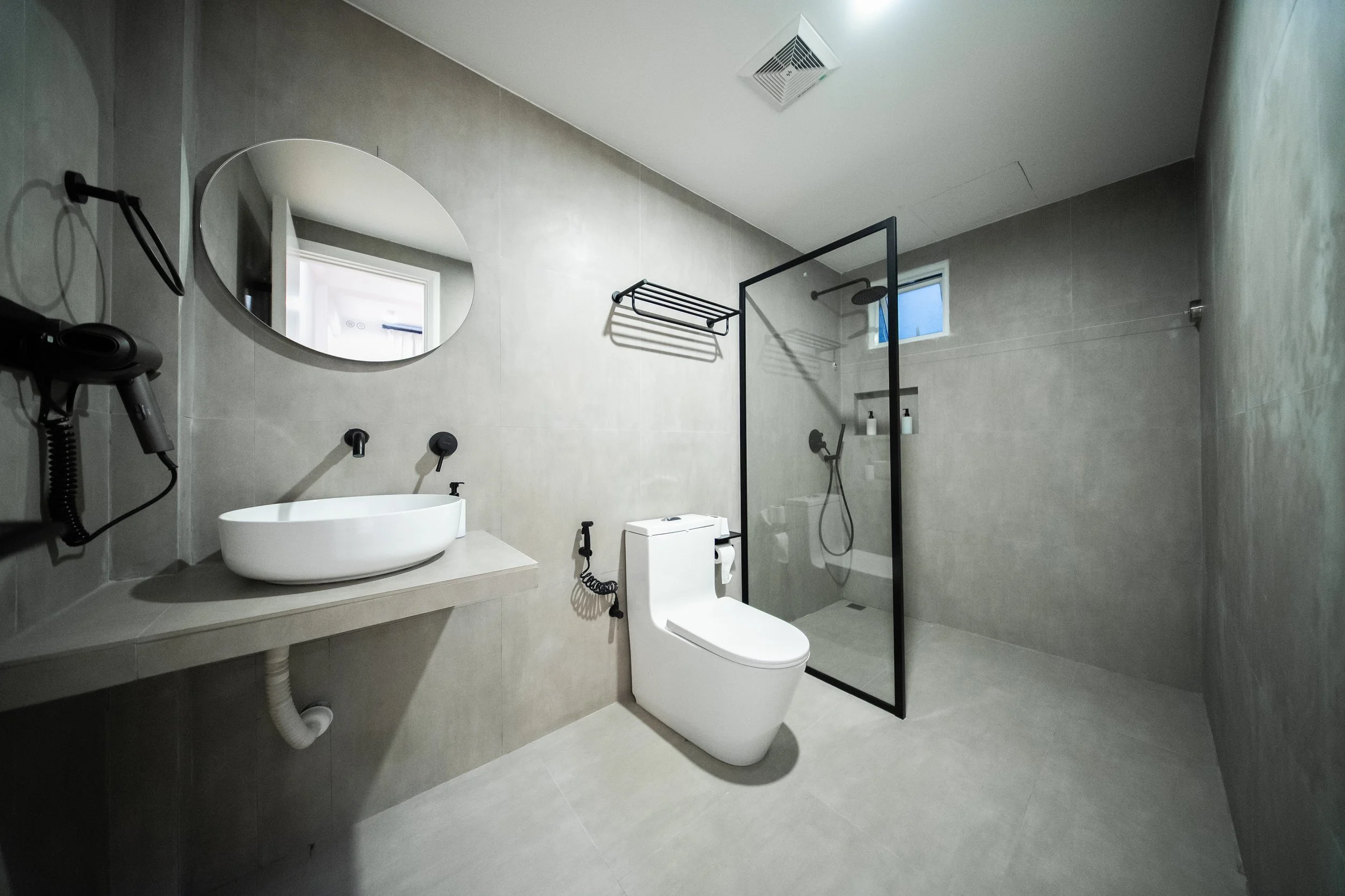 Modern bathroom with a white toilet, wall-mounted sink, oval mirror, wall-mounted hairdryer, walk-in shower with black glass screen, and towel rack.