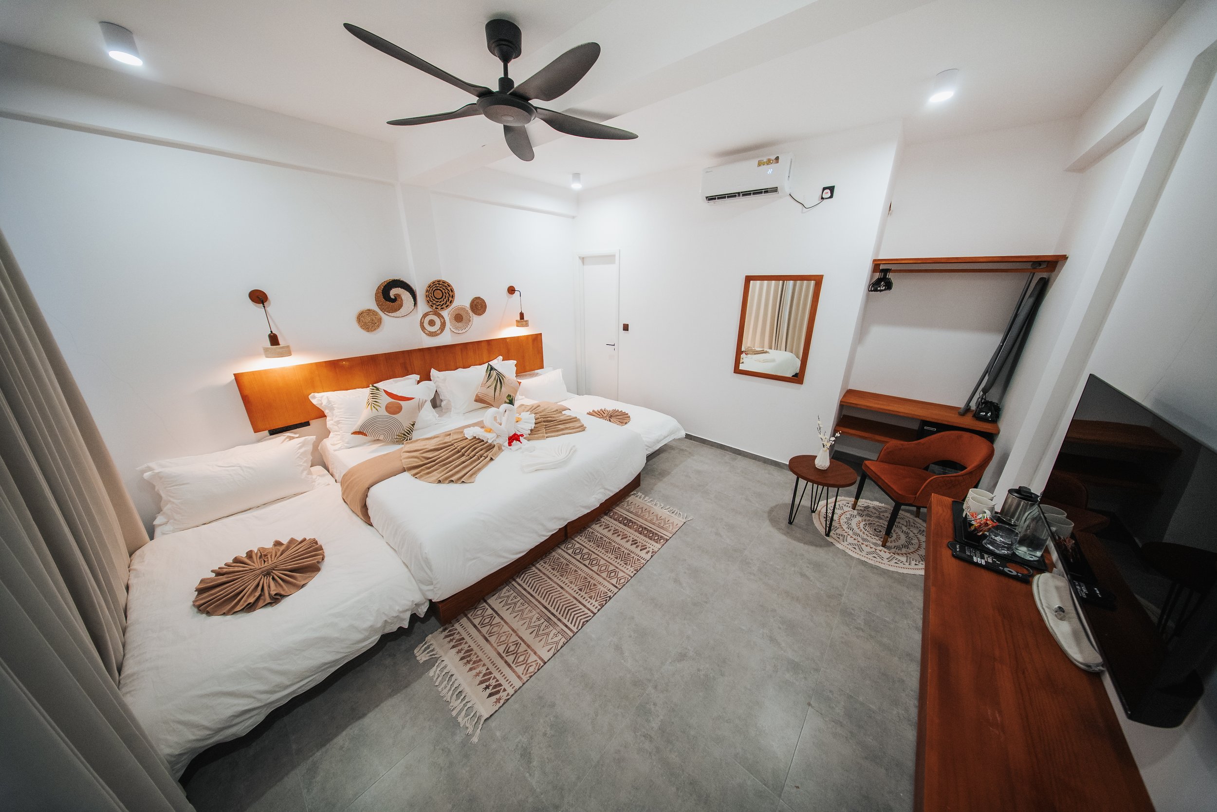 Modern hotel room with a large bed, decorative pillows, and towel sculptures. Room features a ceiling fan, air conditioning unit, wall art, and minimalistic furniture including a chair, side tables, and a desk with a mirror.