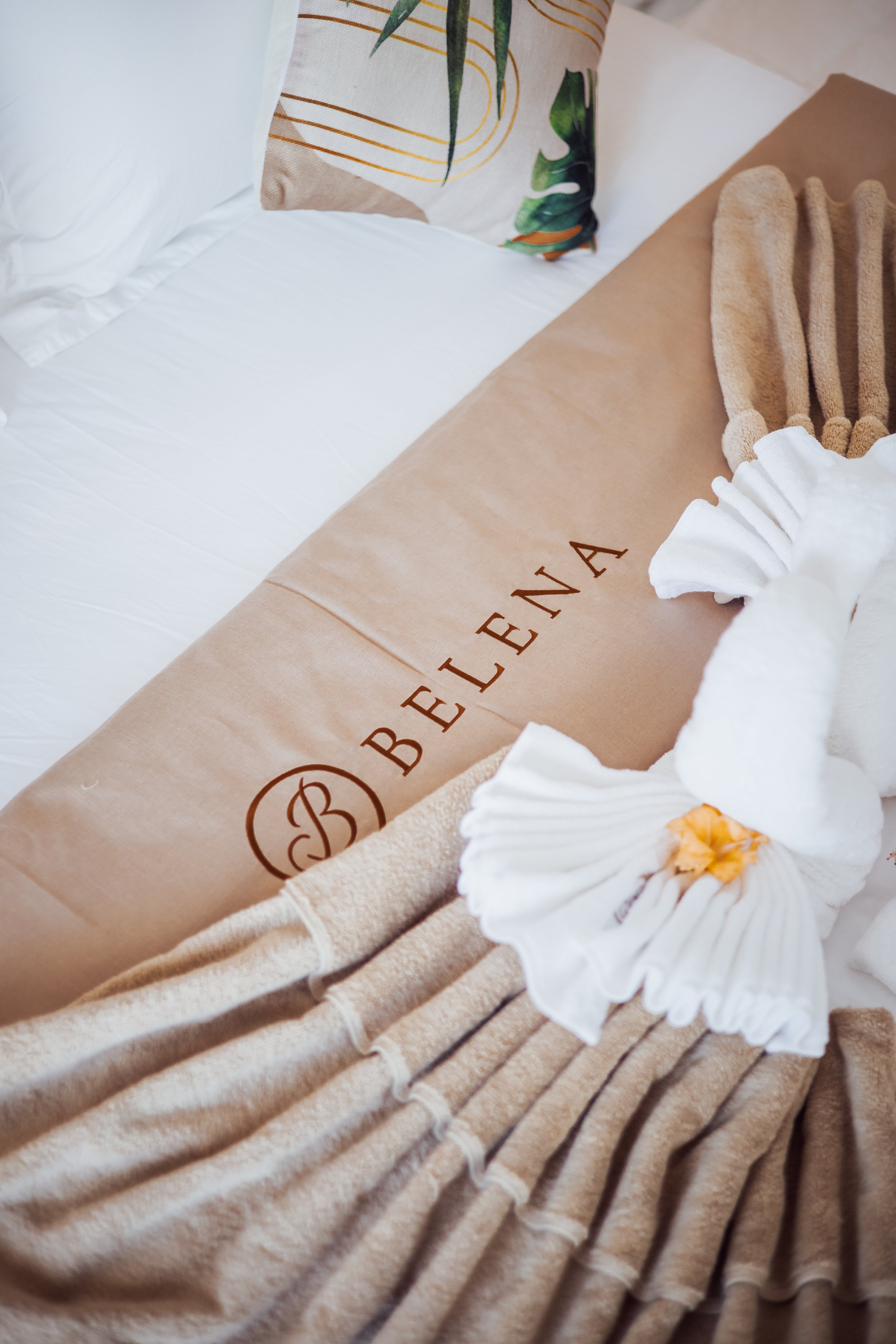 Hotel room bed with decorative towel arrangement and pillow, beige blanket with "Belena" logo.