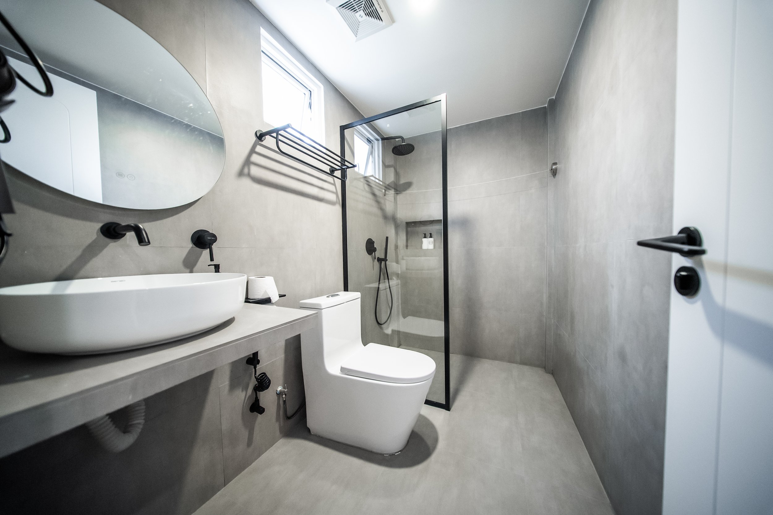Modern bathroom with gray tiles, white toilet, vessel sink, oval mirror, black fixtures, and a glass-enclosed shower.