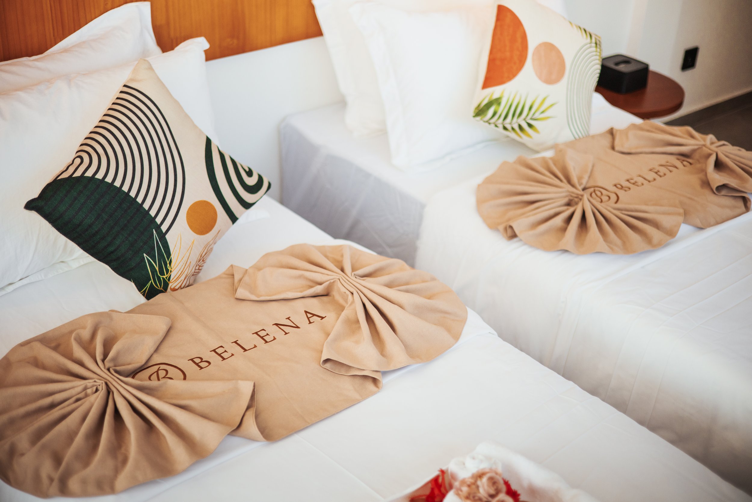 Two neatly made beds with decorative geometric pillows and towels folded with the word 'BELENA' embossed on them.