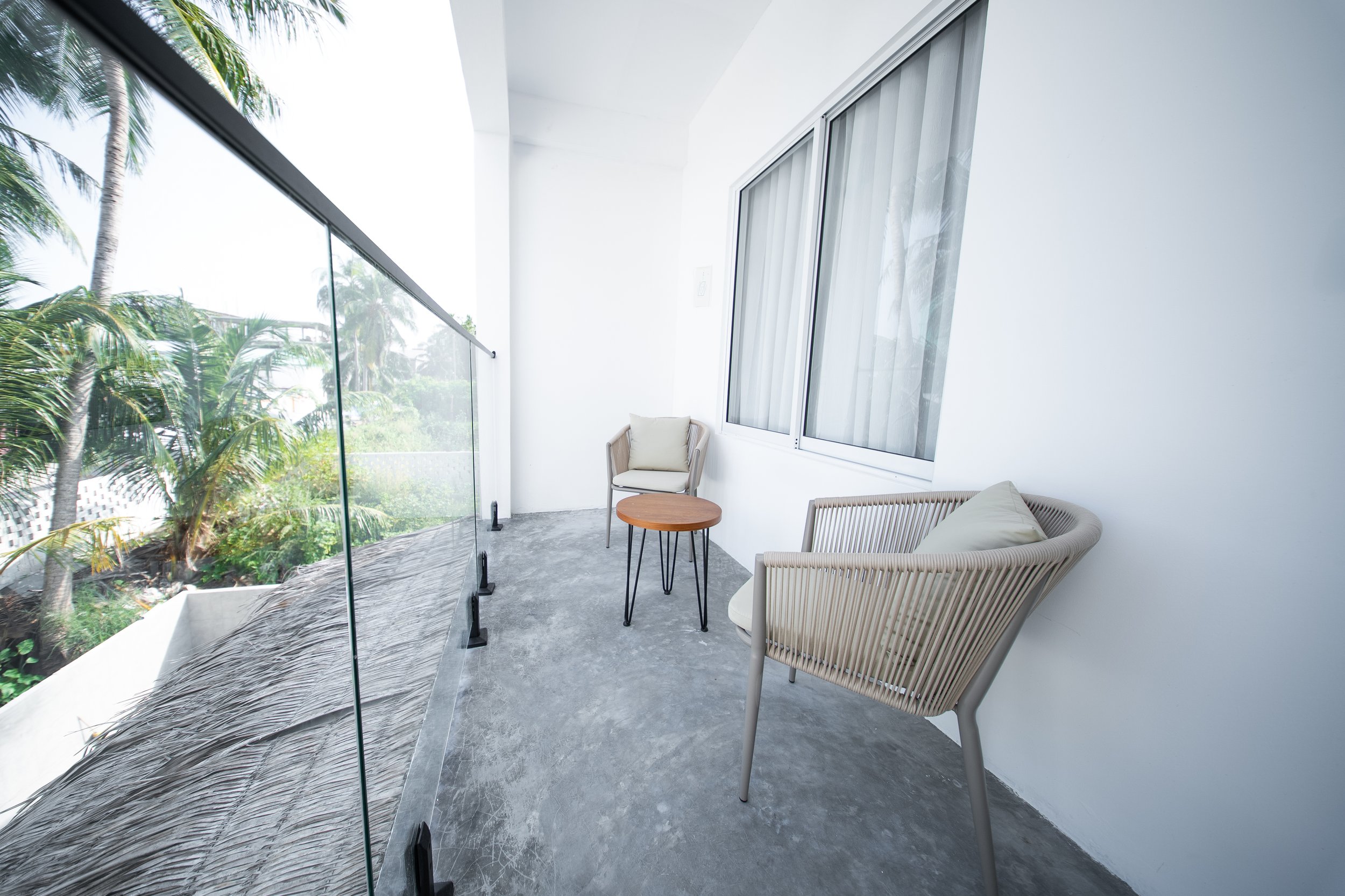 Modern balcony with two wicker chairs, a small round table, glass railing, and tropical plants in the background.