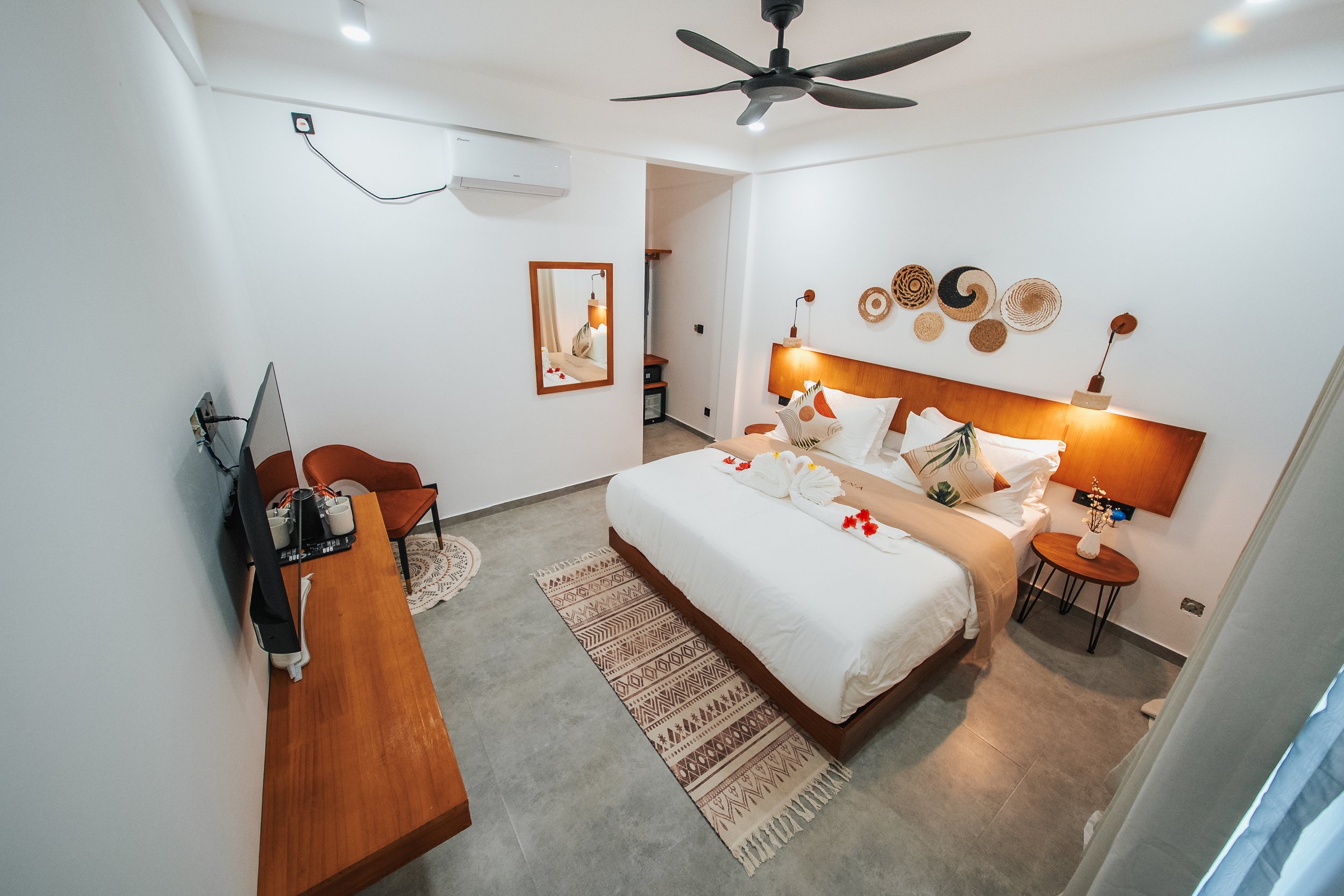 Modern bedroom with a large bed, decorative pillows, and wall art. A ceiling fan hangs above, with a television and chair against the wall. A mirror and bedside tables enhance the decor.
