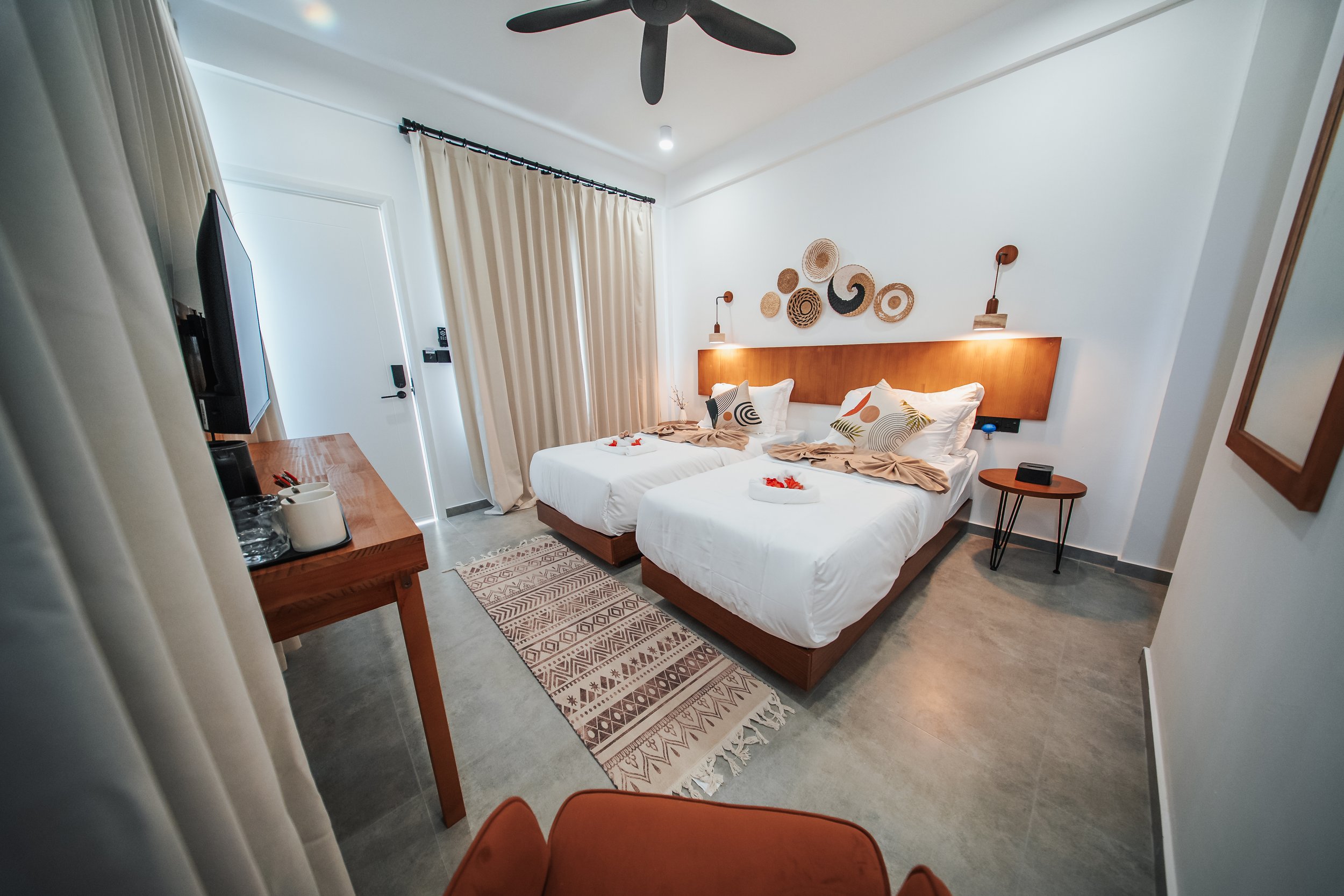 Hotel room with two twin beds, wooden furniture, a ceiling fan, decorative wall art, and a patterned rug. Light curtains cover the window, and there's a TV on a wooden table.