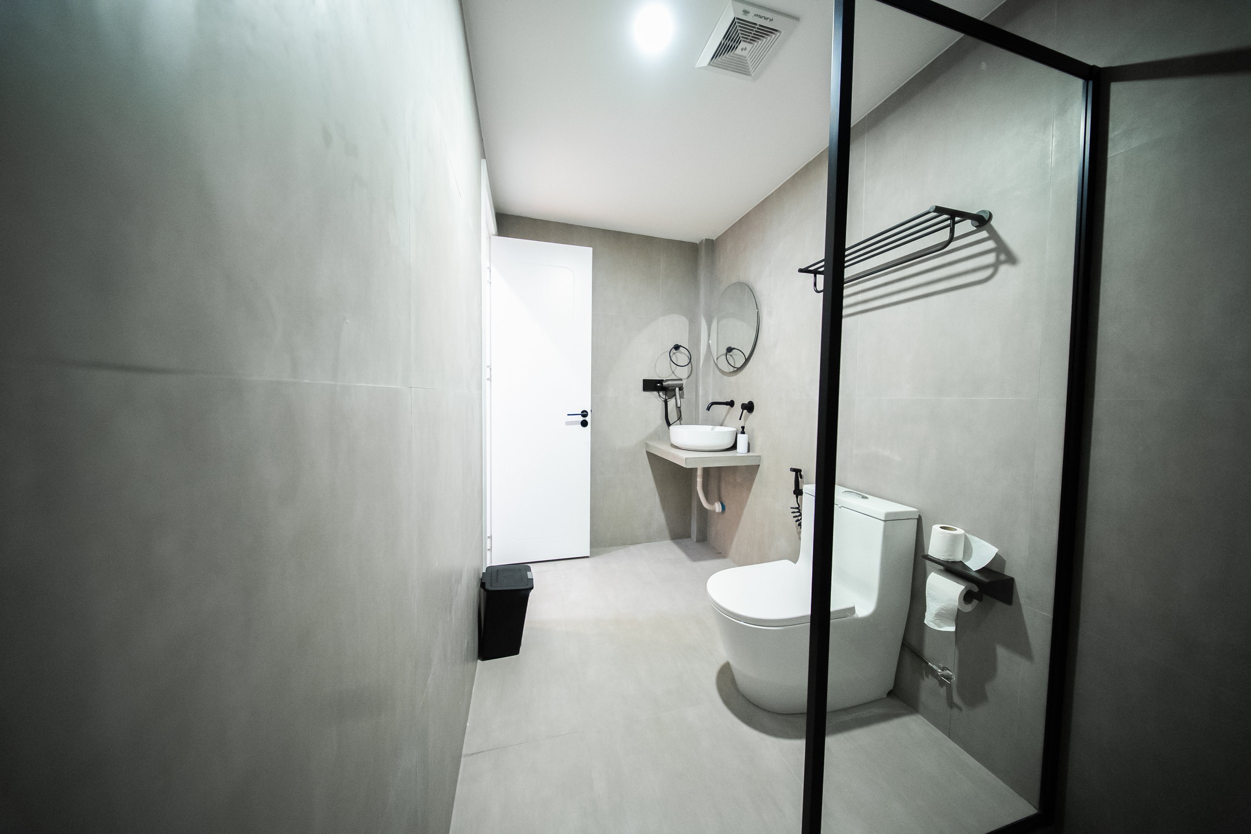Modern minimalist bathroom with gray tile walls, a round mirror above a floating sink, a toilet with a black bidet attachment, towel rack, and a black trash bin.