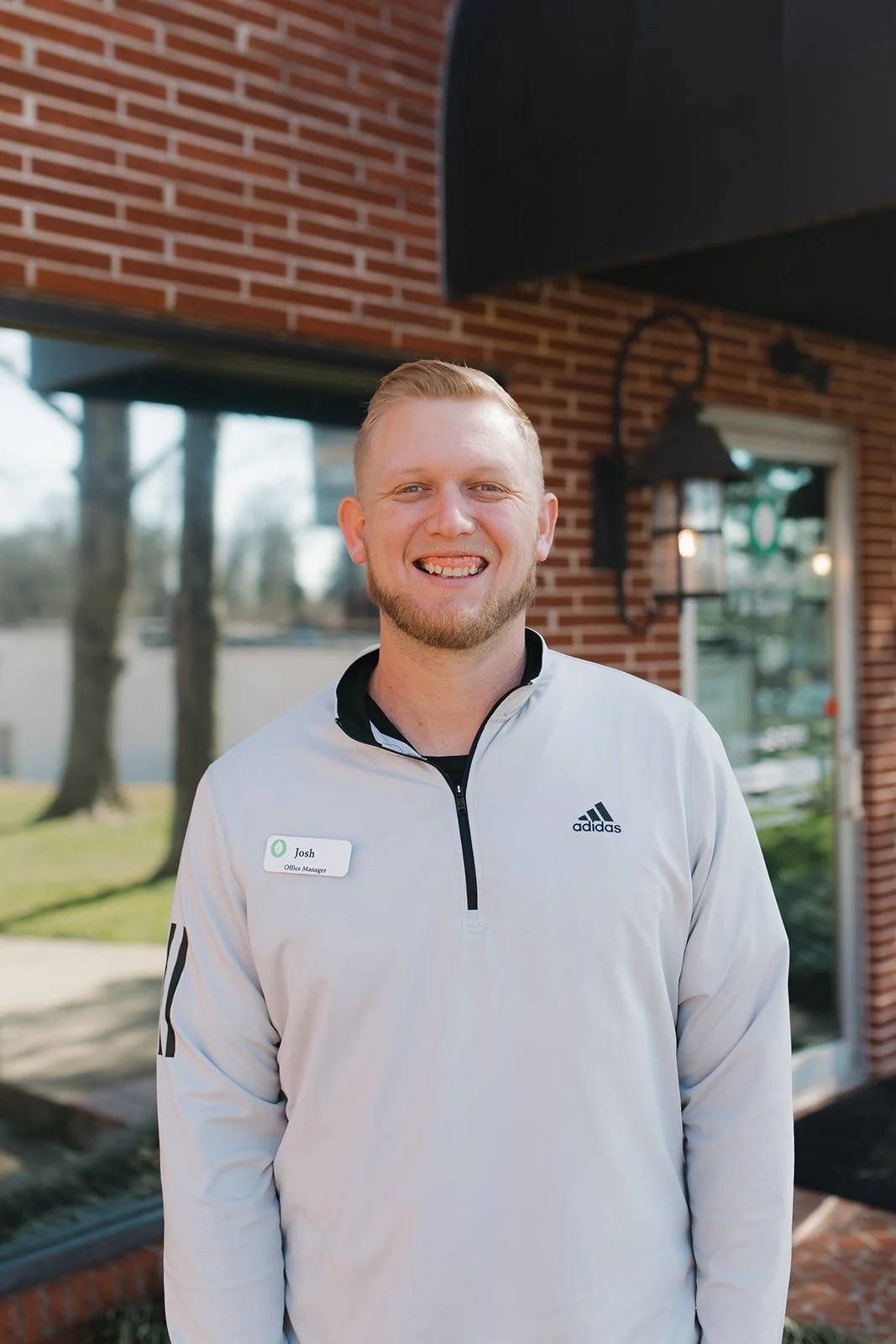 A smiling man wearing a white Adidas pullover with a name tag that says 'Josh Office Manager' standing outside a brick building with large windows and a black outdoor light fixture.