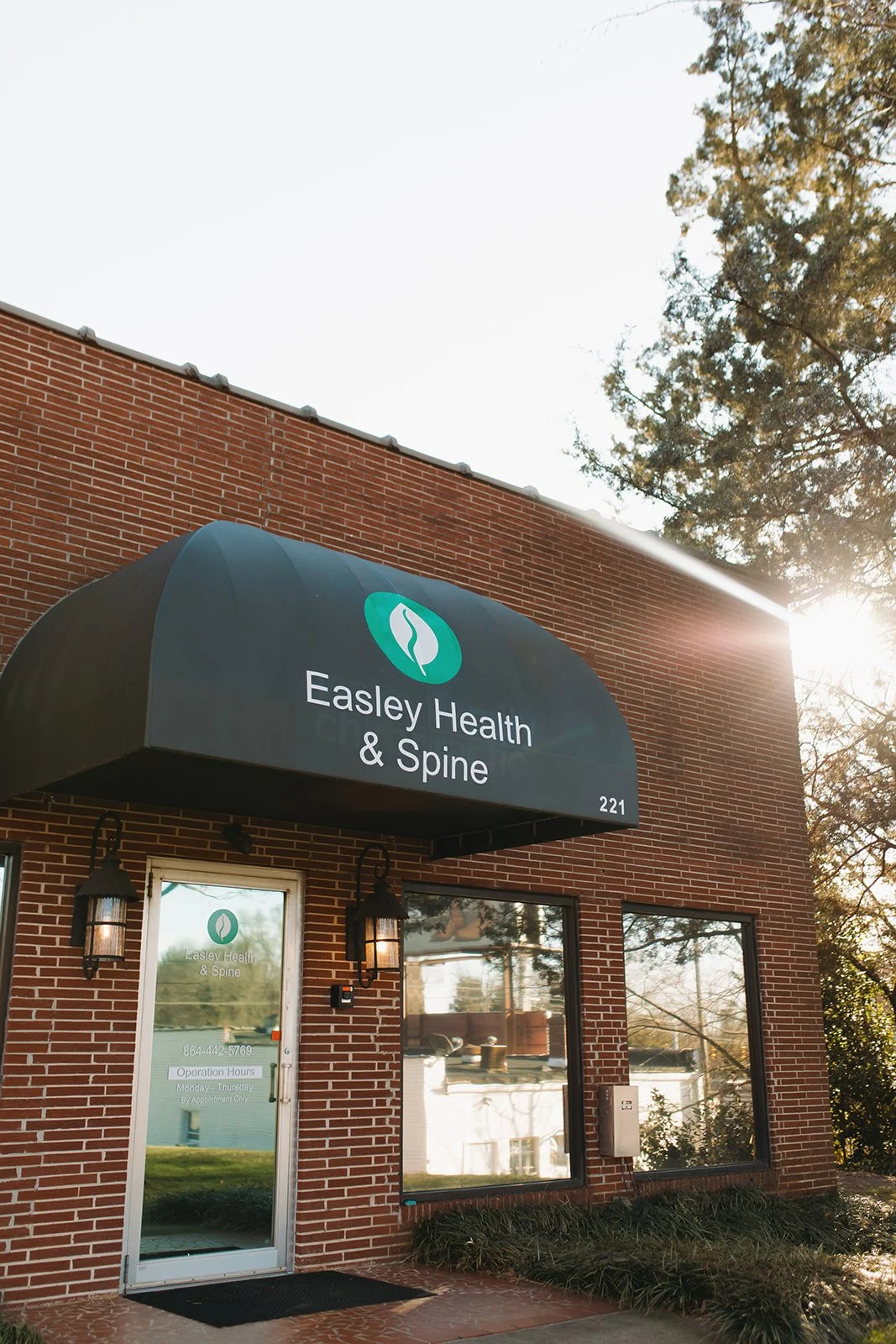 Exterior view of Easley Health & Spine clinic with a brick building, a black awning showing the clinic's name and logo, glass door, and windows reflecting trees and sky, illuminated by sunlight.