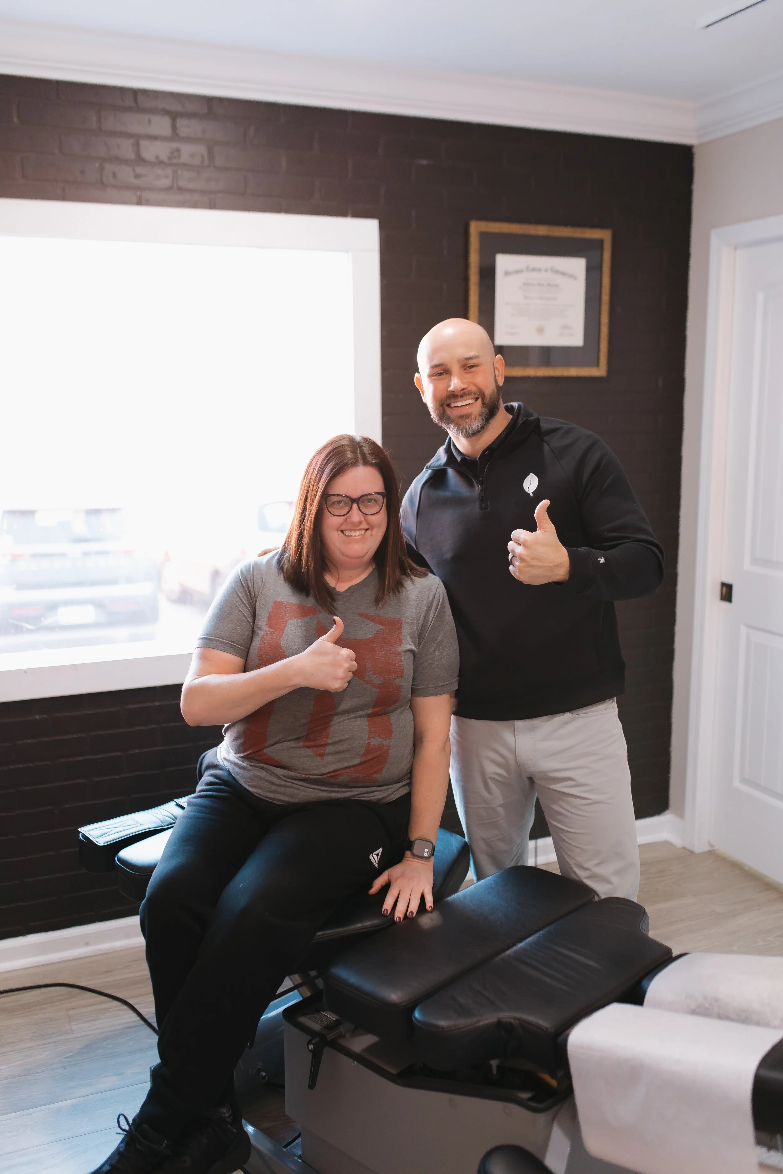 A woman sitting on a treatment table giving a thumbs-up next to a smiling man giving a thumbs-up in a room with a dark brick wall and a picture frame on the wall.