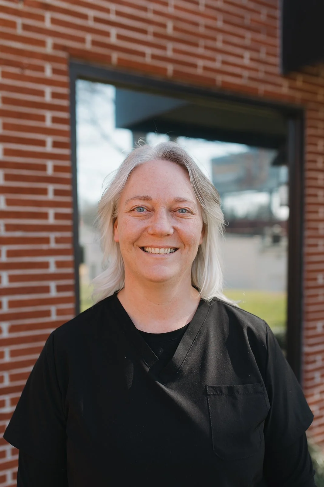 Smiling woman with blonde hair wearing a black shirt standing outdoors in front of a brick building with a large window.