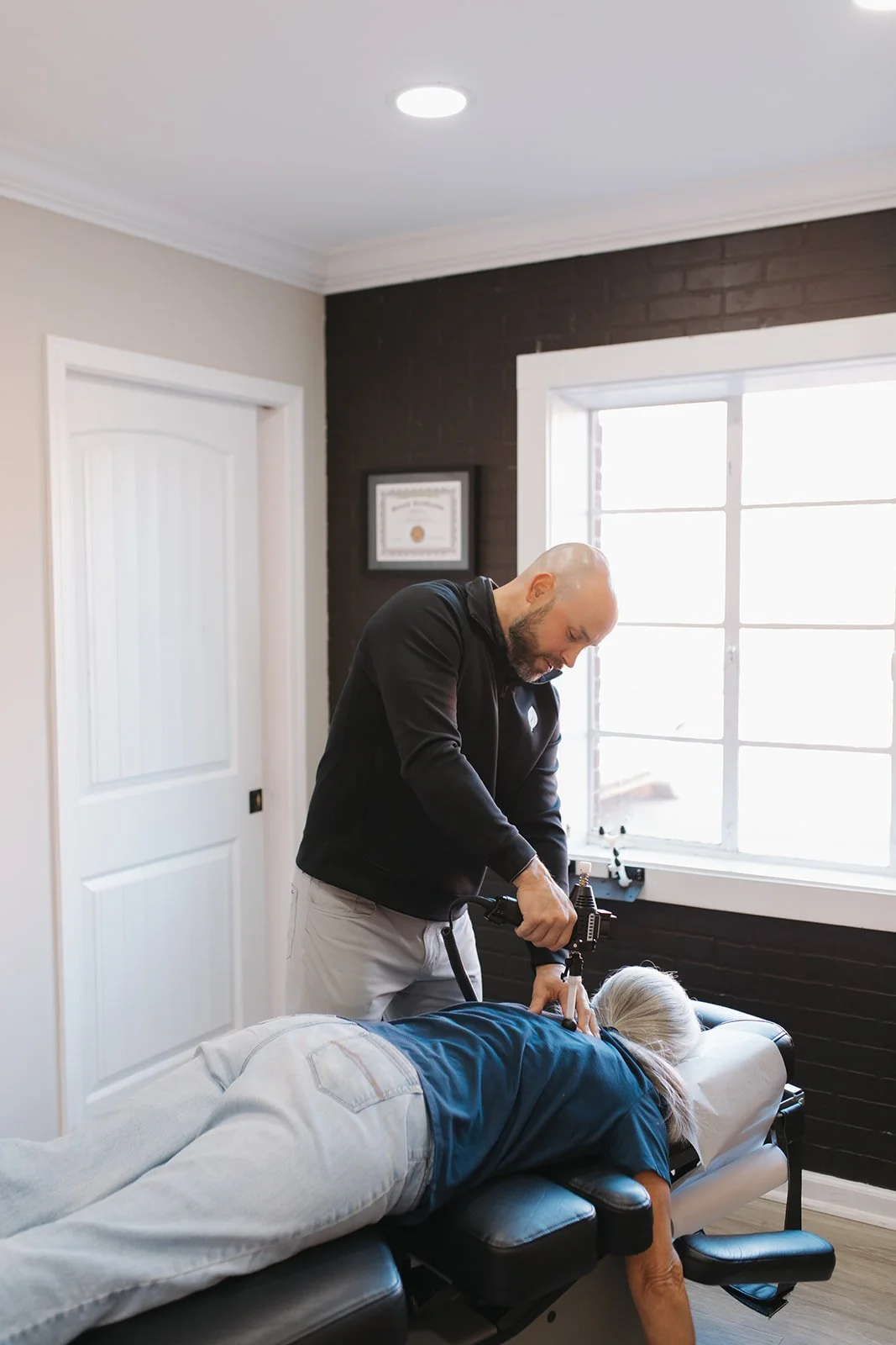 A chiropractor adjusting a female patient lying face down on a chiropractic table in a well-lit room with a window and framed certificate.