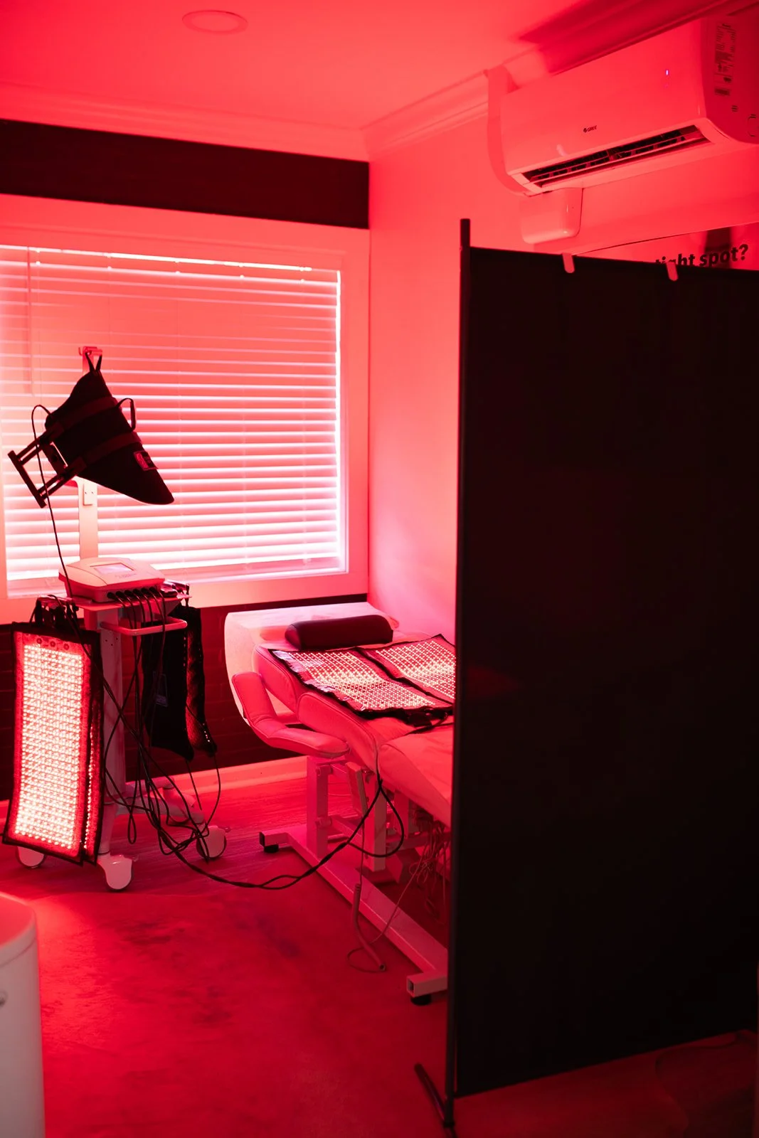 Red light therapy treatment room with red lighting, a treatment bed with pillows, equipment with wires, a window with blinds, and an air conditioning unit.