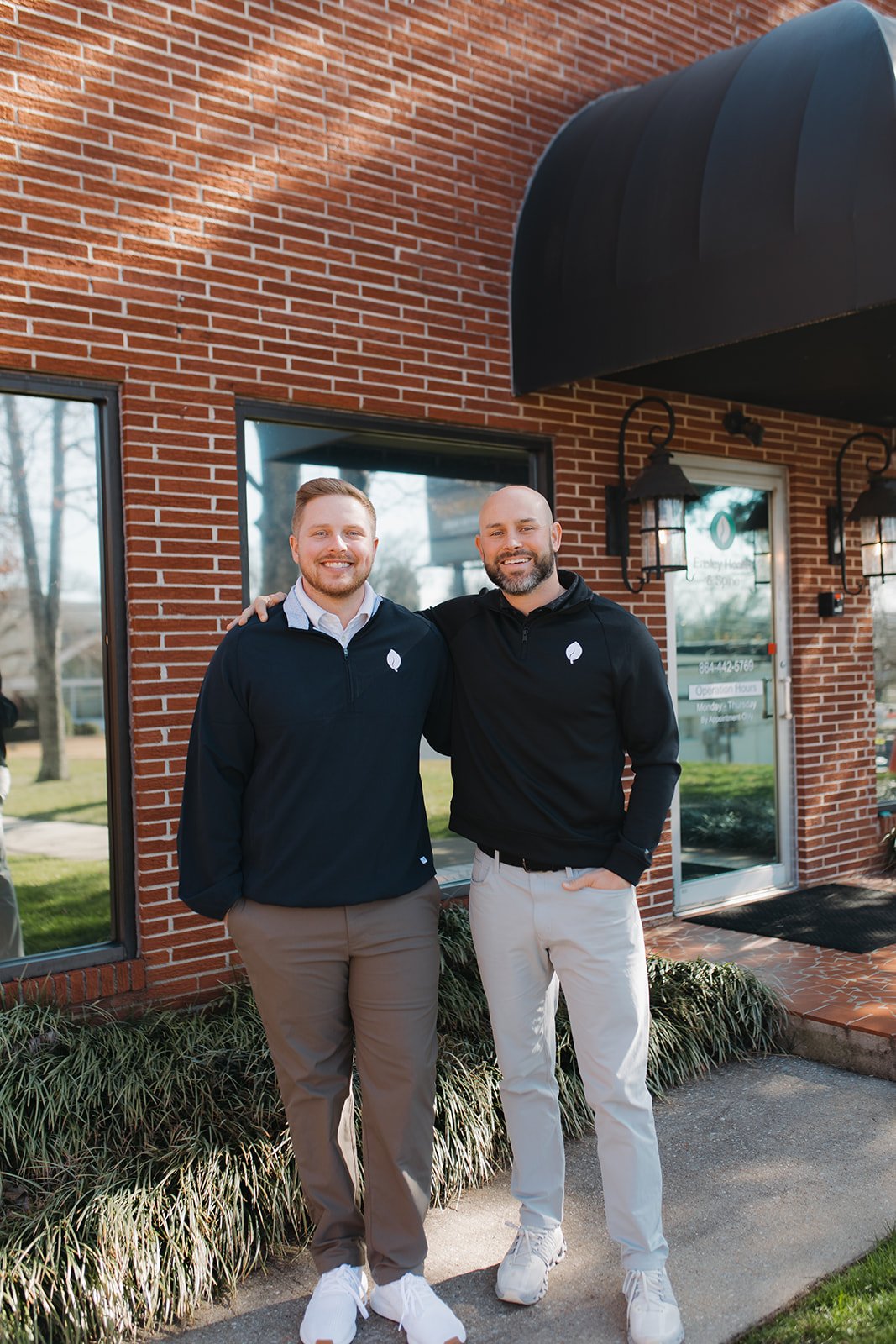 Two men standing side by side outside a brick building with large windows, smiling. One has his arm around the other's shoulder. Both are wearing black jackets with white leaf logos, beige pants, and white sneakers. The building has black awnings and outdoor wall lanterns.