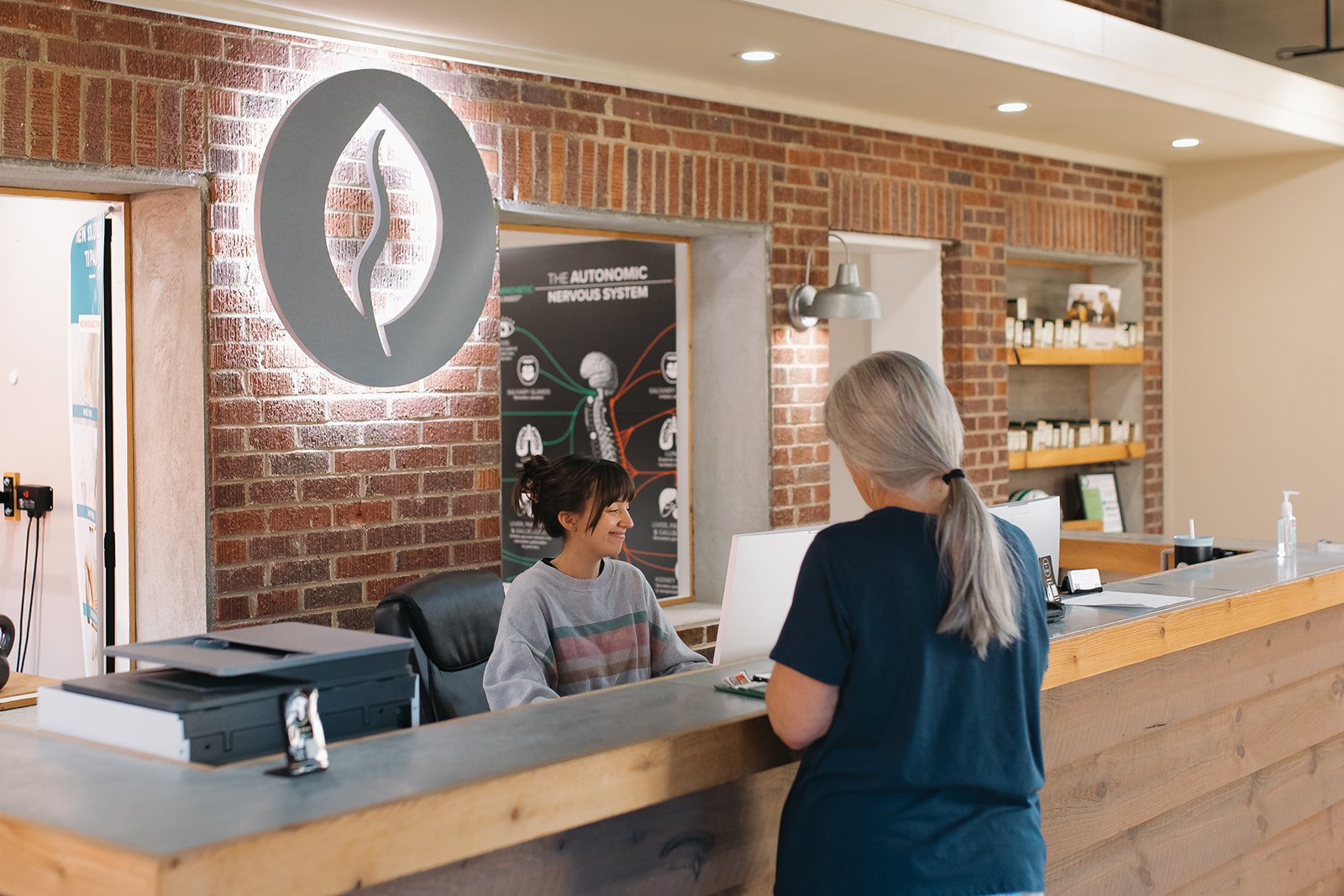 A woman with gray hair and a blue shirt checking in at a reception desk to a woman with short dark hair wearing a gray sweatshirt, inside a building with exposed brick walls and a logo of a leaf or plant on the wall behind the woman at the desk.