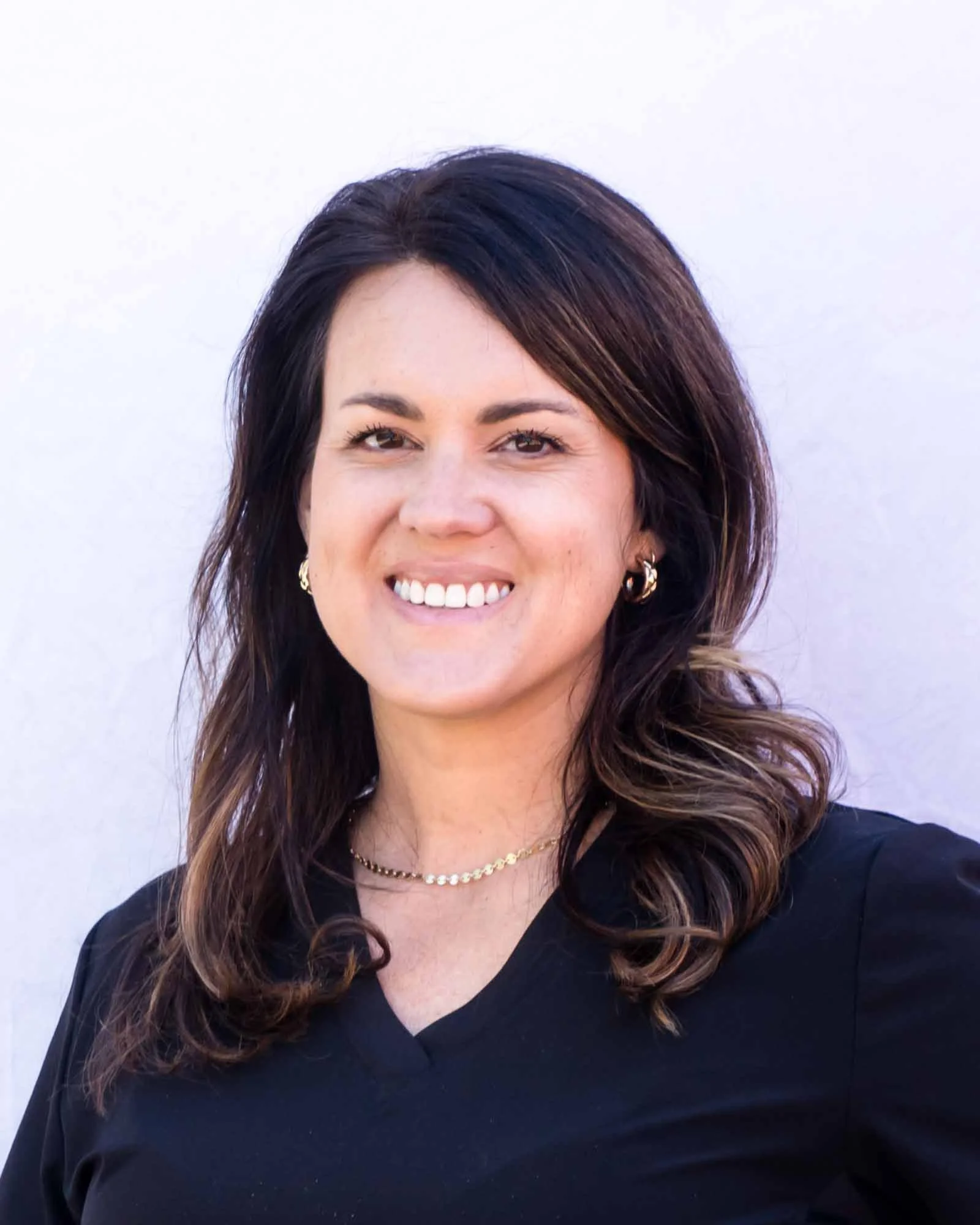A nurse with shoulder-length brown hair smiling, wearing a black top, gold jewelry, and standing against a plain light background.
