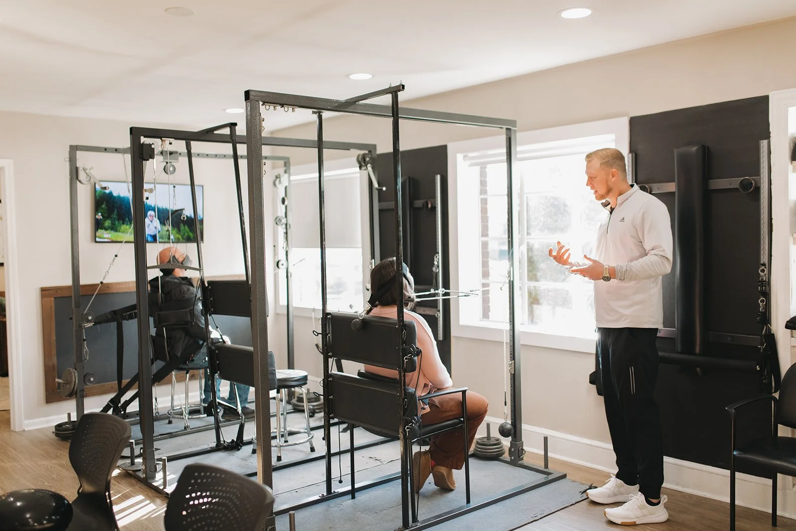 A chiropractic traction expert explaining techniques to a woman seated in a specialized traction machine, with another person using the traction machine in the background, inside a well-lit chiropractic office.