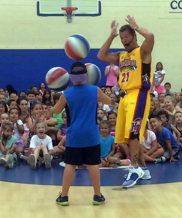 The Hoopwizard is wearing a yellow Los Angeles Lakers jersey with the number 21 interacts with a young boy at a basketball event, with a crowd of children watching excitedly in the background.