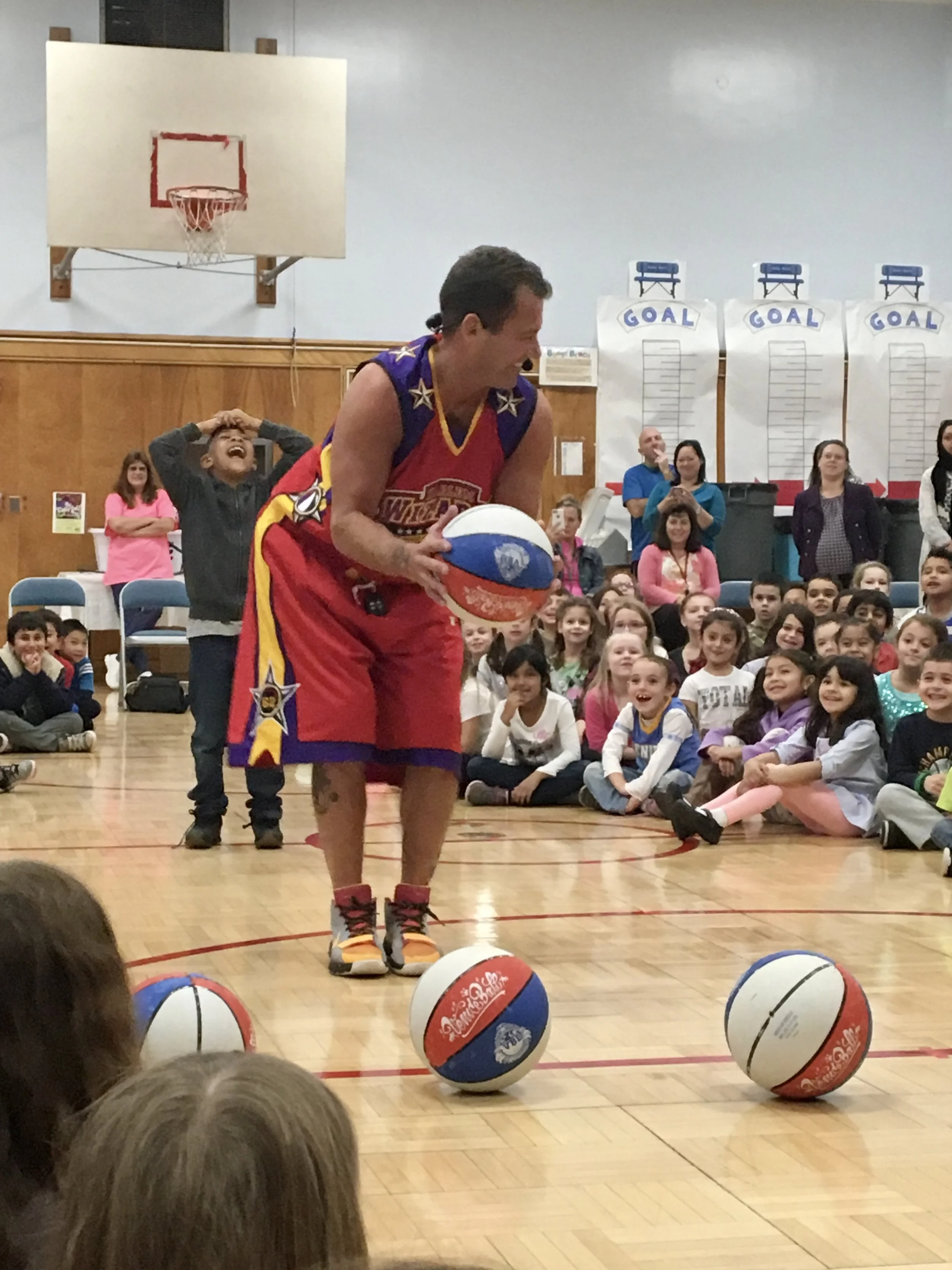 The Hoopwizard is holding a basketball on a gymnasium court, performing a trick for a seated audience of children and adults. There are three more basketballs on the floor, and the kids are smiling and laughing.