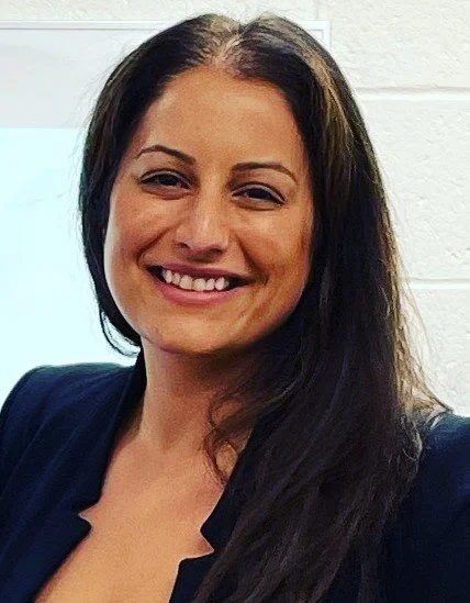 A woman with long dark hair smiling, wearing a dark blazer, in front of a white brick wall.