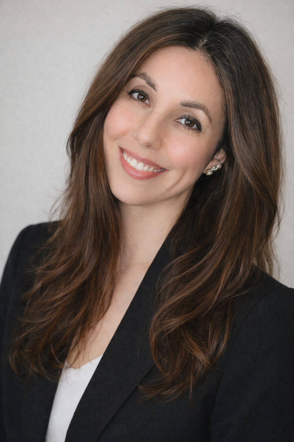 A woman with long dark hair smiling, wearing a dark blazer, in front of a white brick wall.