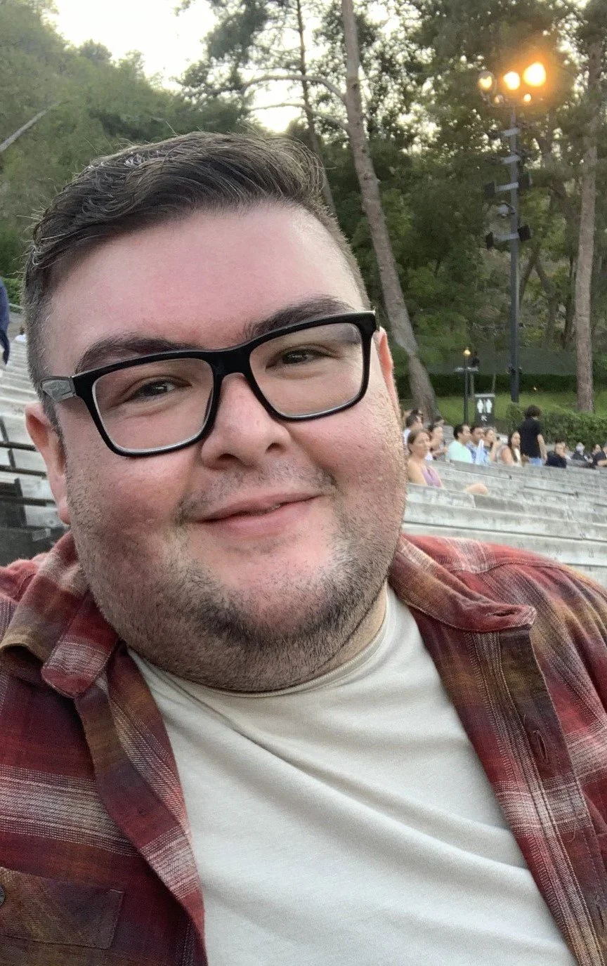 A smiling man wearing glasses, a white t-shirt, and a plaid shirt, sitting outdoors at a stadium or park with trees and a crowd of people in the background during evening lighting.