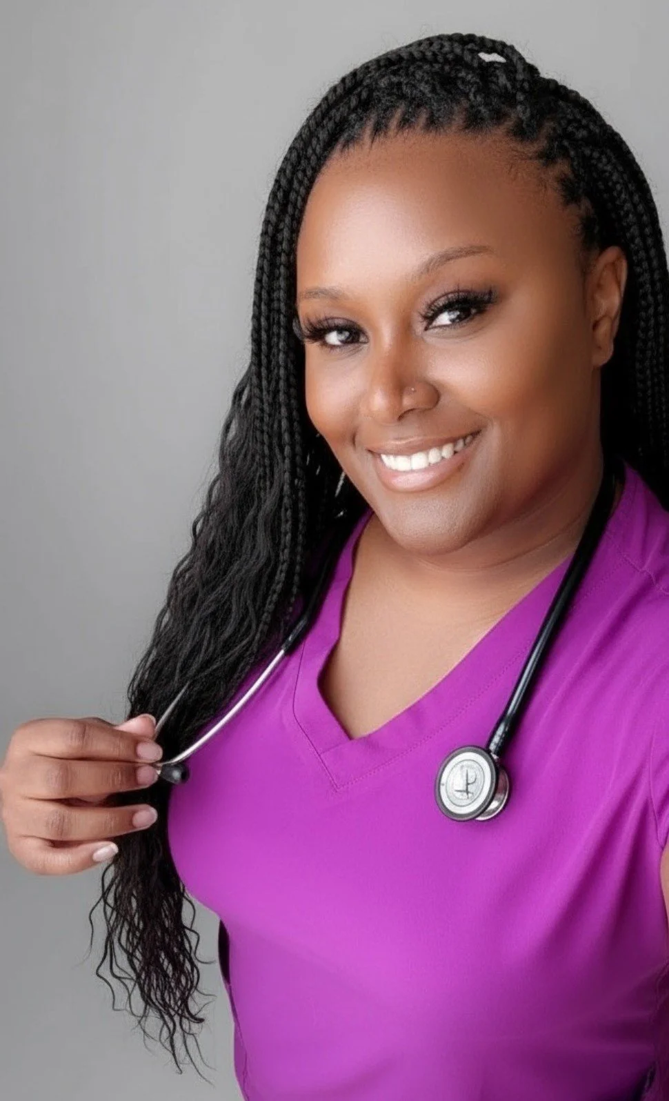 A female healthcare worker smiling at the camera, wearing black scrubs and a stethoscope, standing in a medical setting.