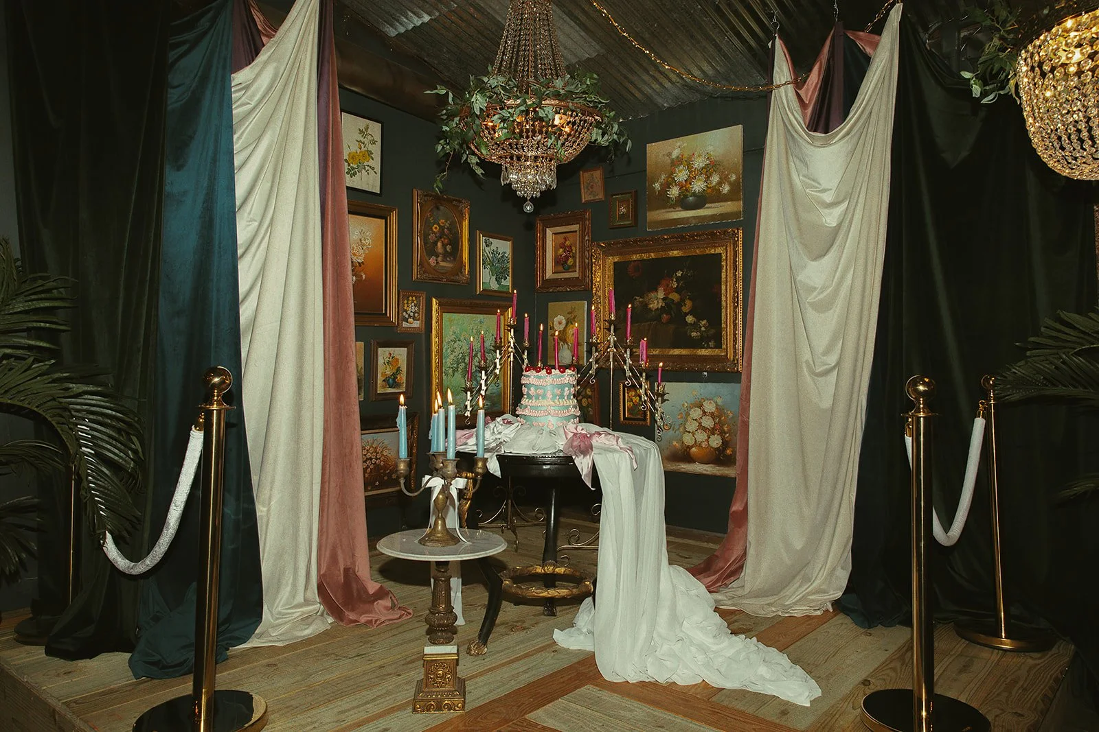 An art gallery themed setup featuring paintings on dark-colored walls, draped curtains in blue, pink, and cream, and a chandelier with greenery. At the center, there is an antique table with a white tablecloth, topped with a pink frosted cake, pink candles, and candelabras with lit blue and pink candles, surrounded by gold stanchions with white ropes.