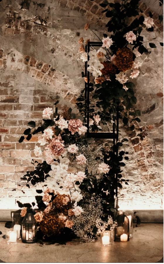 Floral arrangement with pink, white, and brown flowers against an exposed brick wall, illuminated by candles on the ground.