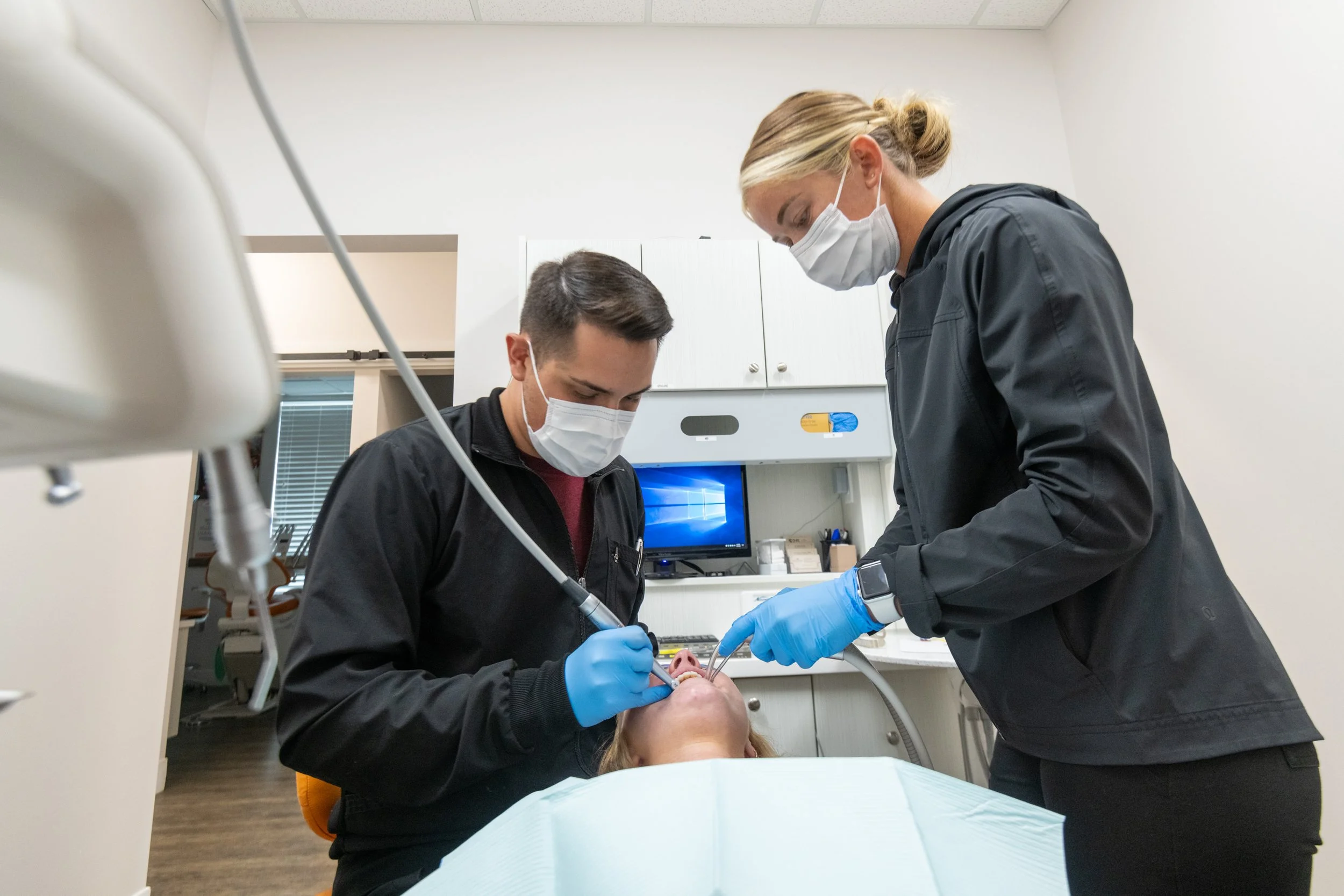 Two dental professionals wearing masks and gloves performing a dental procedure on a patient lying in a dental chair in a clinical office.