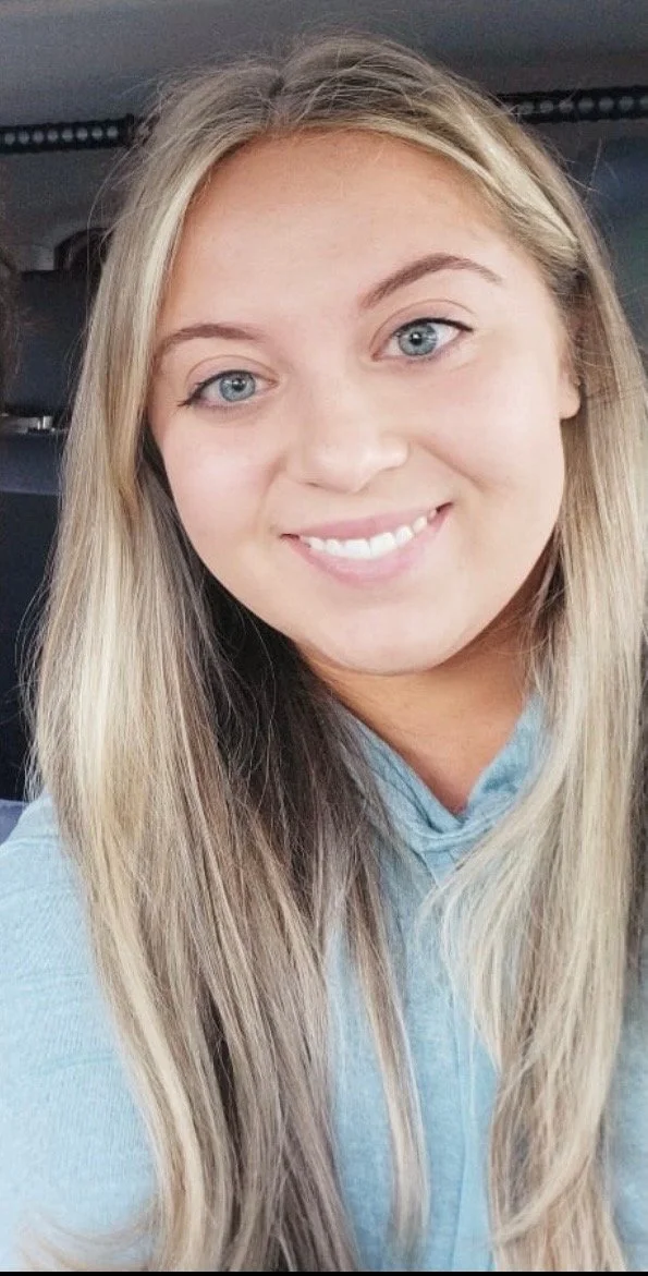 Close-up of a smiling young woman with long blonde hair and blue eyes, wearing a light blue hoodie.