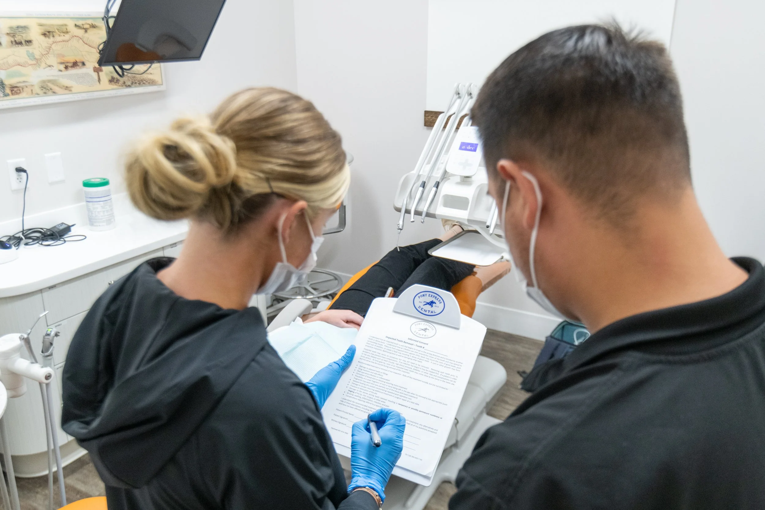 Dental clinic with two dental professionals reviewing a document, patient lying on dental chair, dental tools on the wall, and a monitor on the wall.