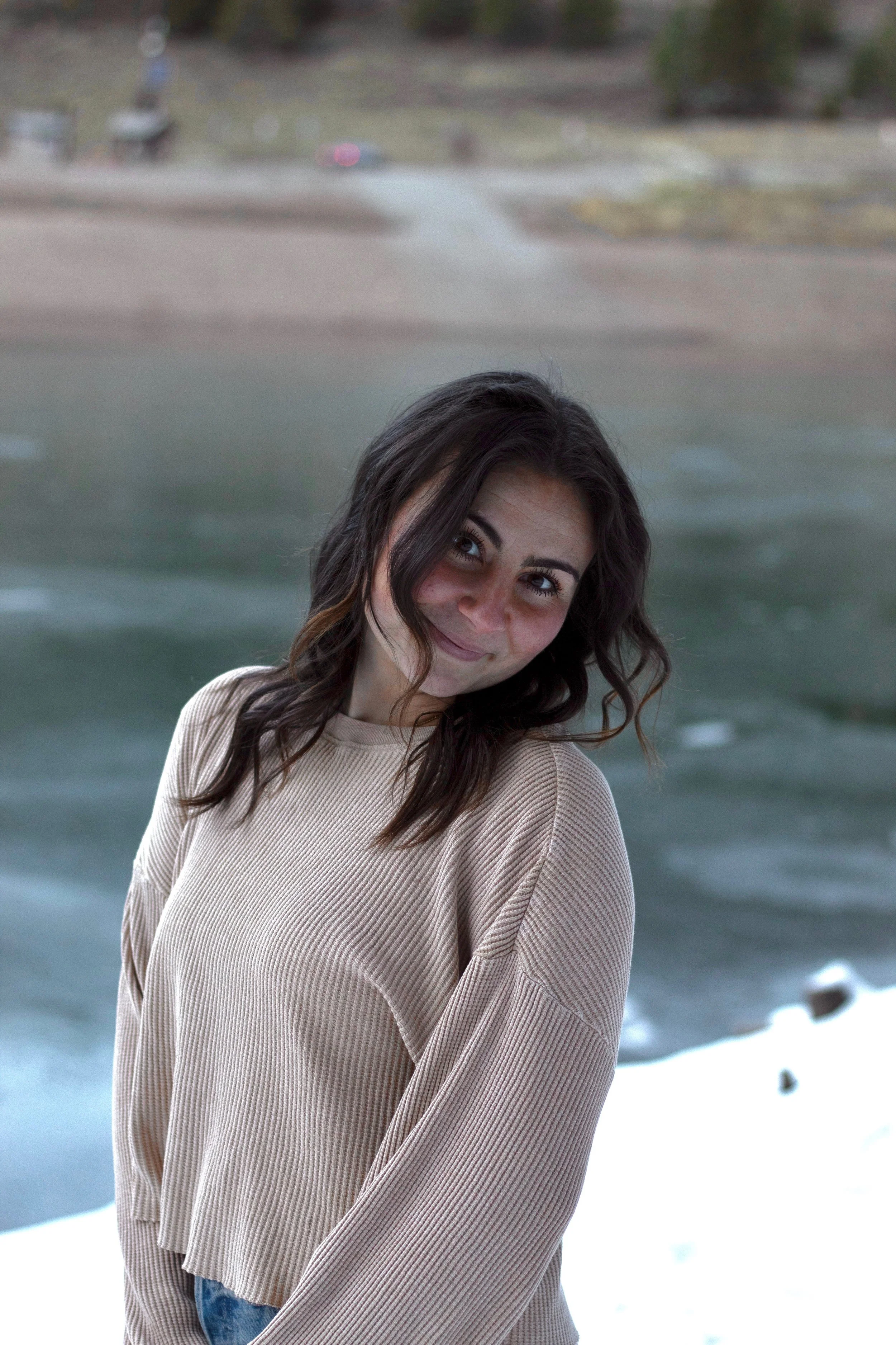 Young woman with dark wavy hair smiling outdoors near a river with snow on the ground.