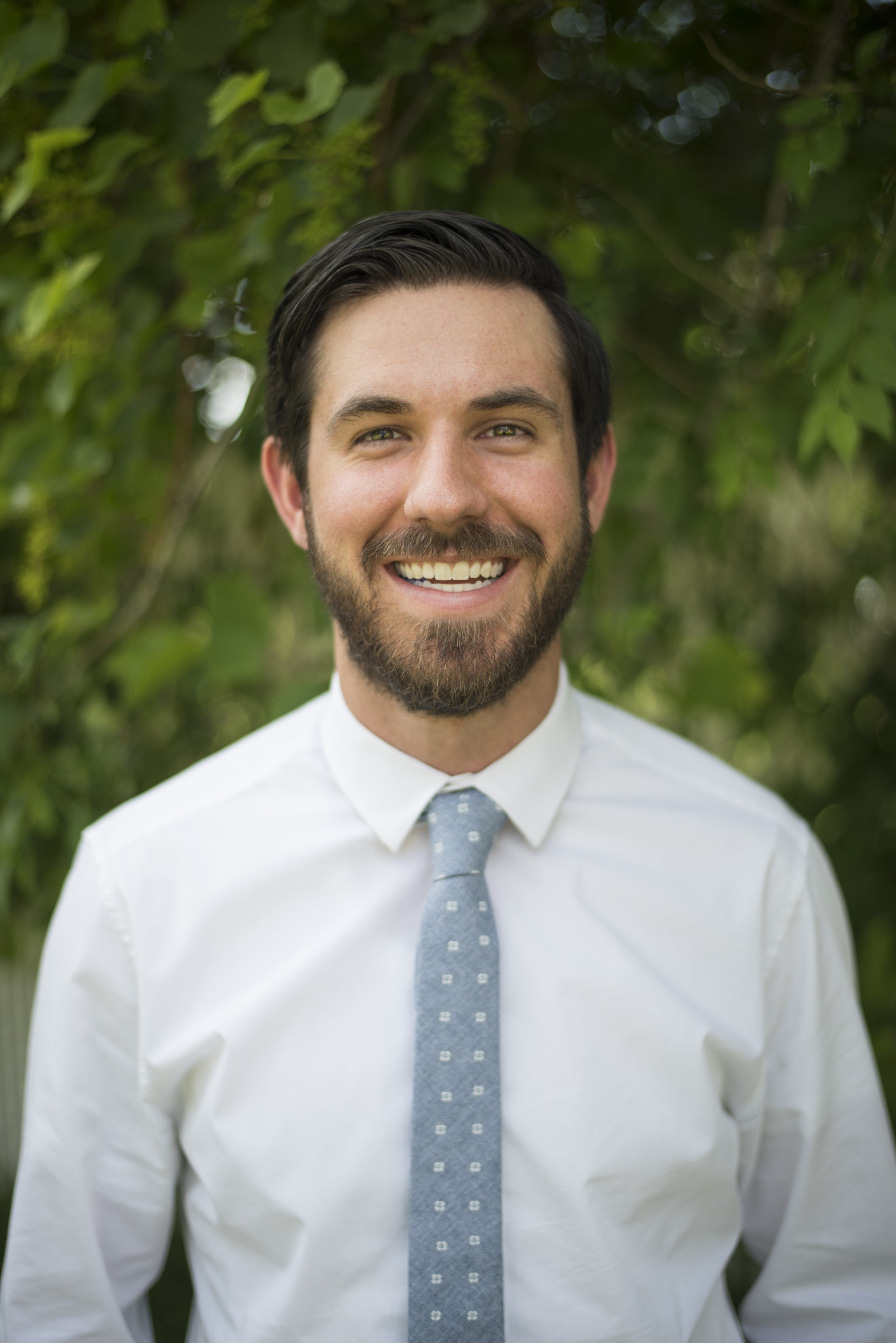 A smiling man with dark hair and a beard, wearing a white shirt and a light grey tie, standing outdoors with green foliage in the background.