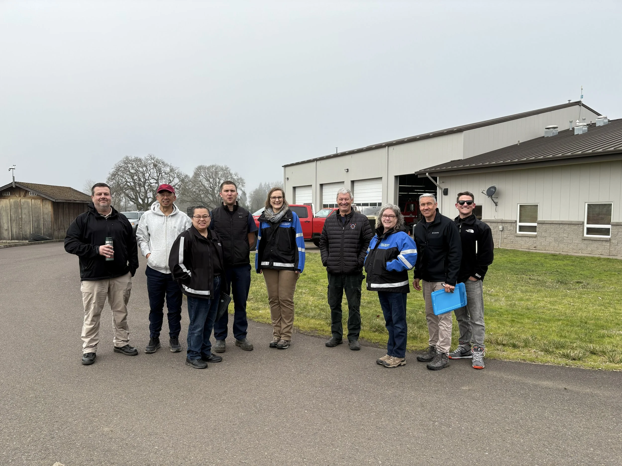 Group of nine people standing outside in front of a metal building, smiling and posing for the photo.