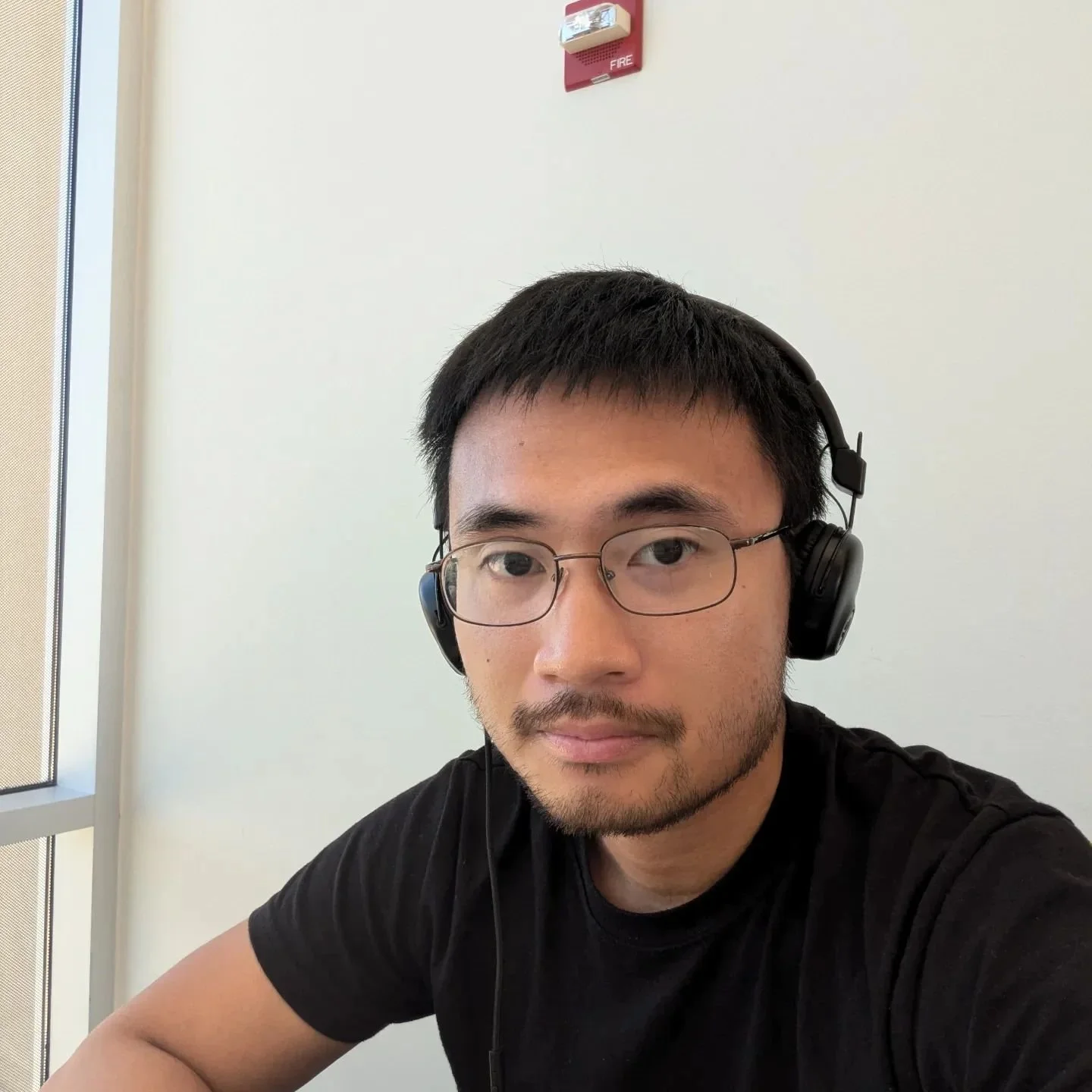 A young man with black hair, glasses, and a beard wearing headphones and a black t-shirt, sitting indoors near a window with a white wall and a red fire alarm above him.
