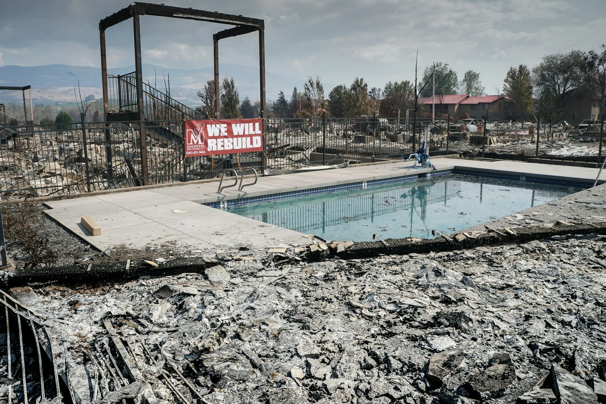 Photograph showing the aftermath of a wildfire with a community pool surrounded by rubble and sign saying “We Will Rebuild”