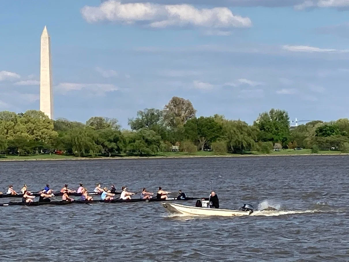 Daytime photograph showing two boats of rowers practicing on the Potomac River with a coach supervising from a motorboat and the Washington Monument in the background