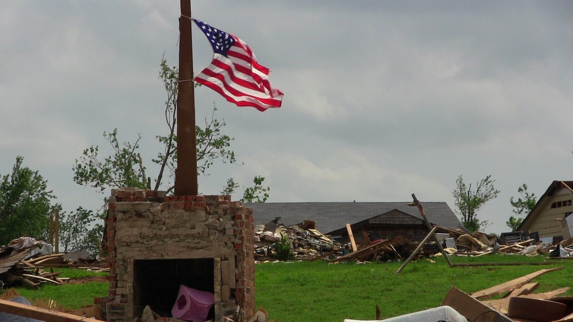 Photo of a destroyed home with additional rubble in the background and a US flag flying in the foreground