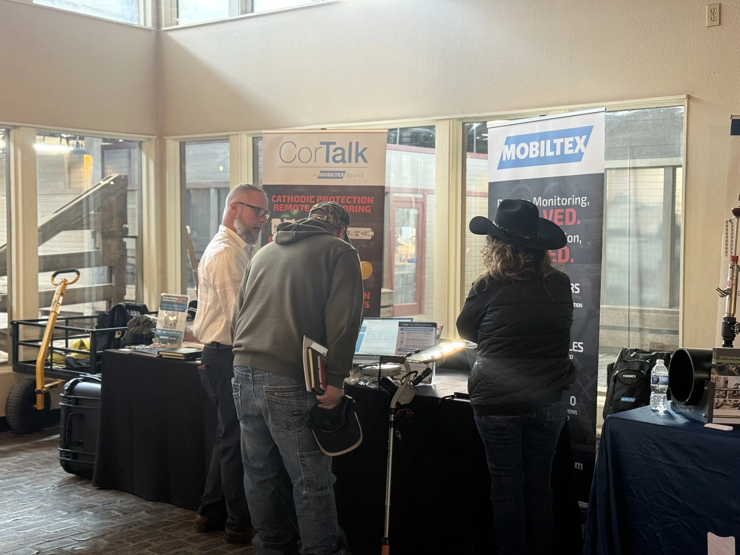 Three people at a booth with banners for CorTalk and MobileX, inside a room with large windowed walls and wooden floors.