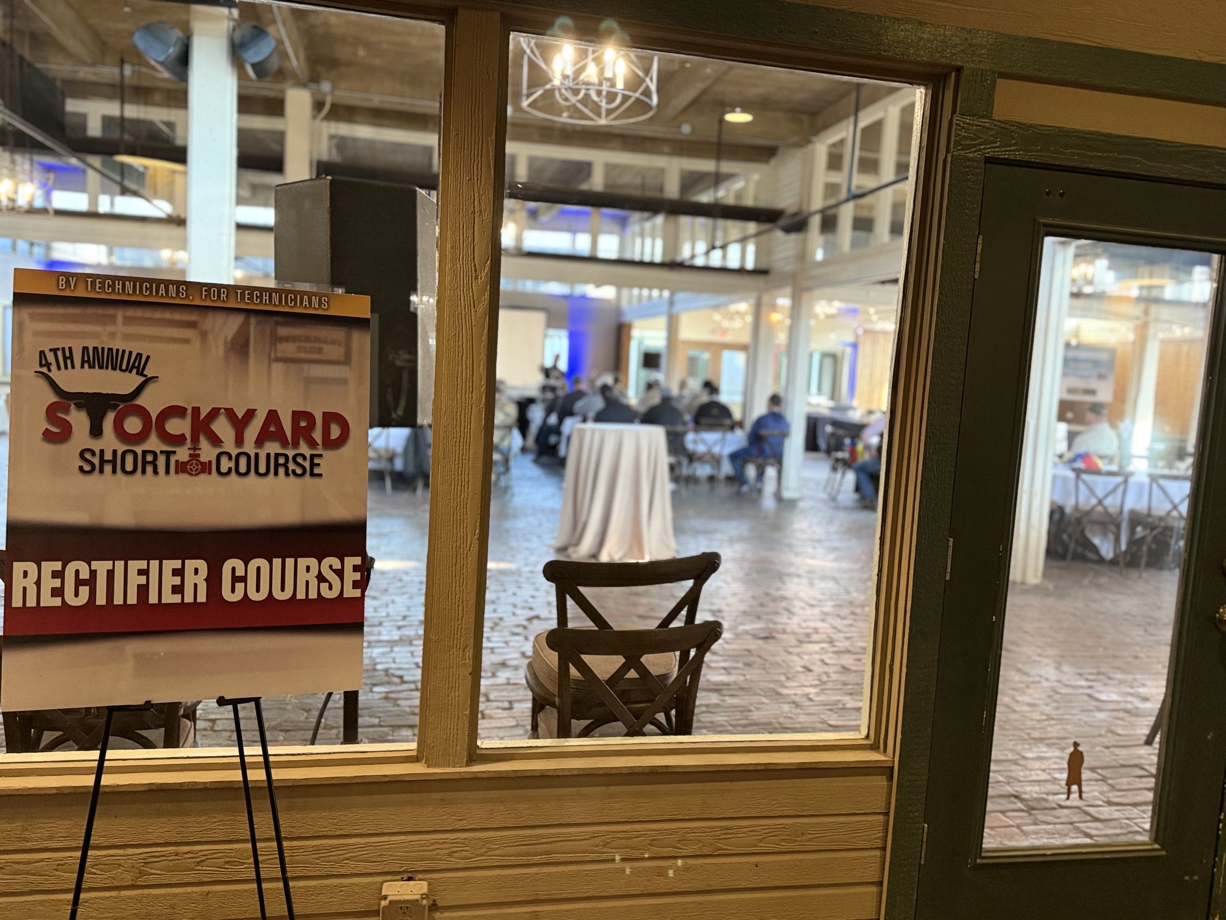 View through a window and door into an indoor seating area at a venue hosting the 4th annual Stockyard Short Course, showing tables with chairs, people seated, and some standing, with industrial-style decor and lighting.