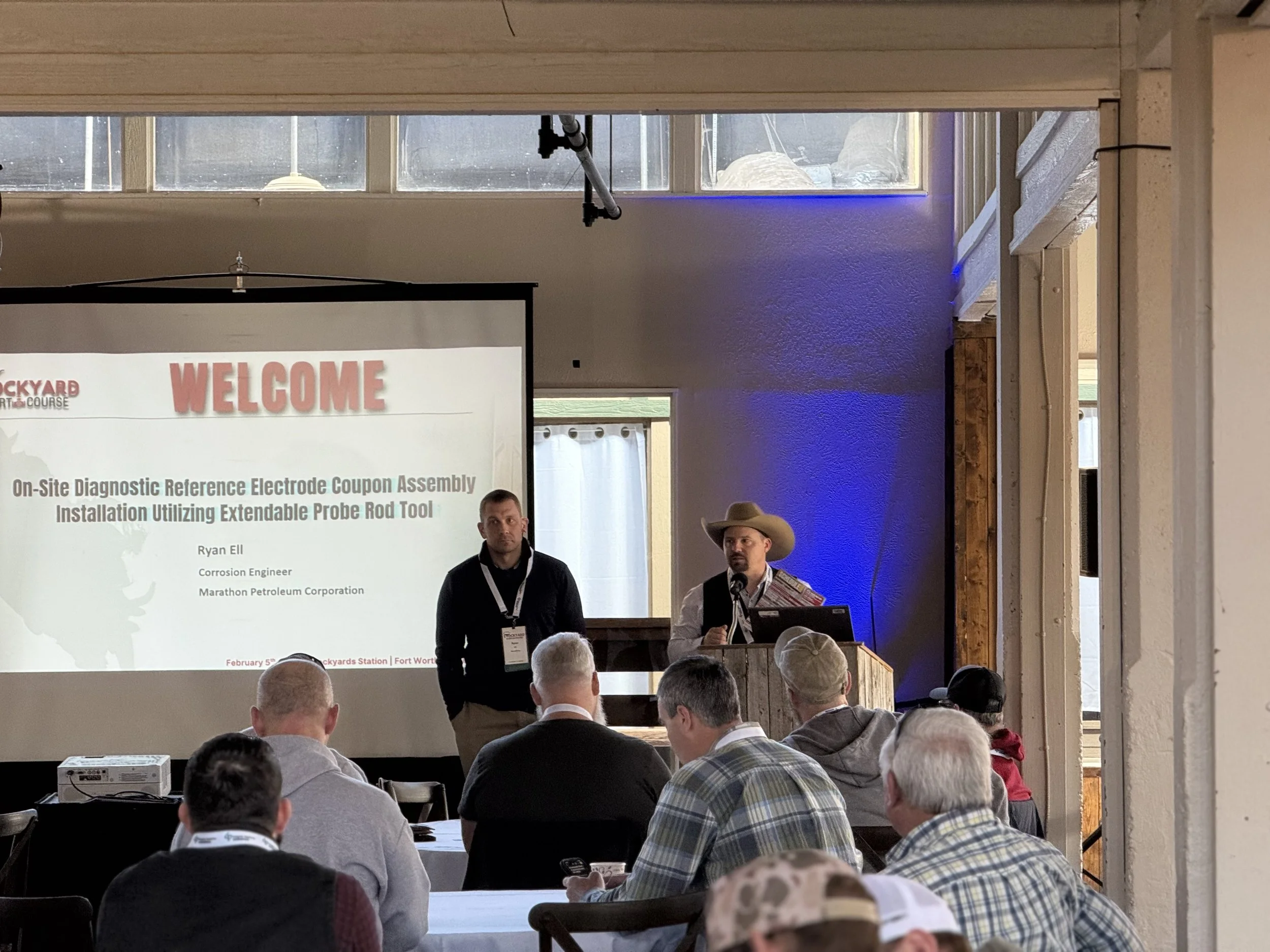 A conference room with people attending a presentation on onsite diagnostic electrode coupon assembly. A large screen displays a welcome message and details about the presentation. Two men are standing near the screen, one speaking into a microphone 