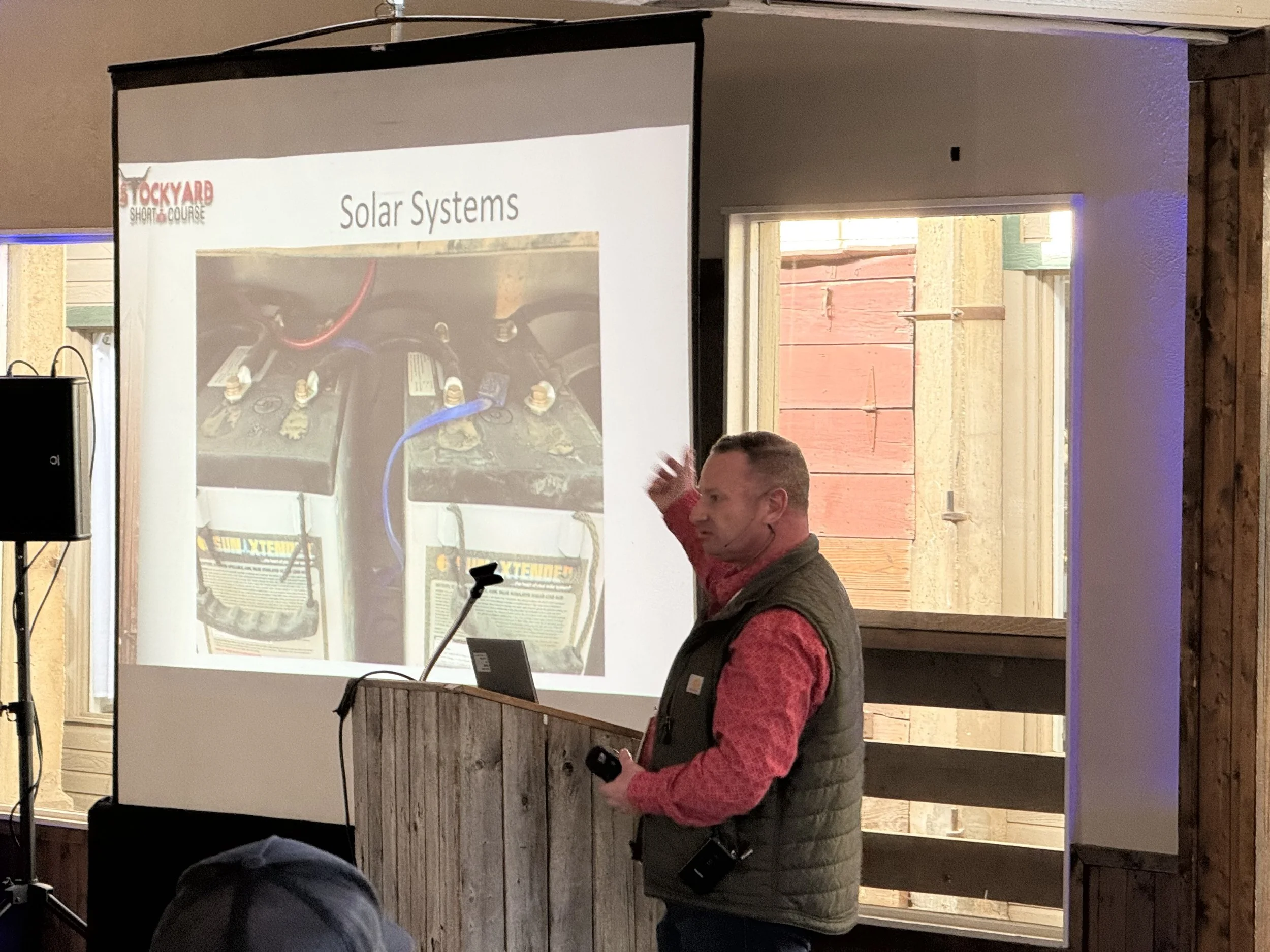A man giving a presentation on solar systems at a wooden podium, with a slide projected on a screen behind him showing batteries connected to a solar system, in a rustic room with wooden walls and windows.