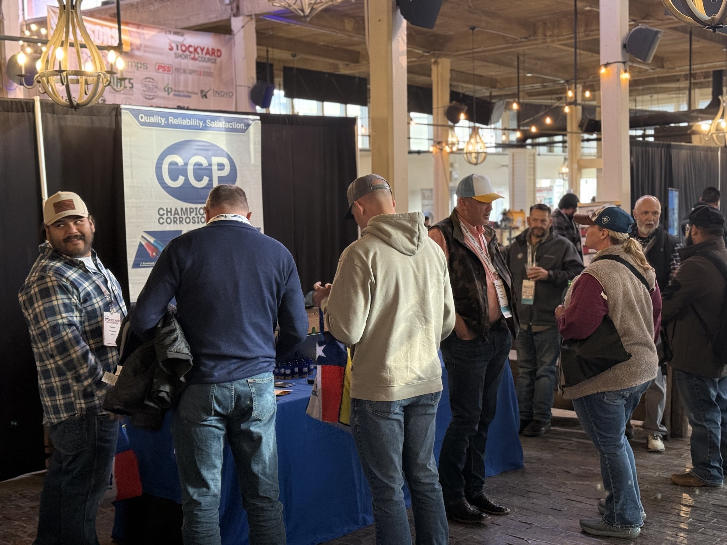 People gathered around a booth at an indoor event, with banners and display tables, in a venue with wooden beams and string lights.