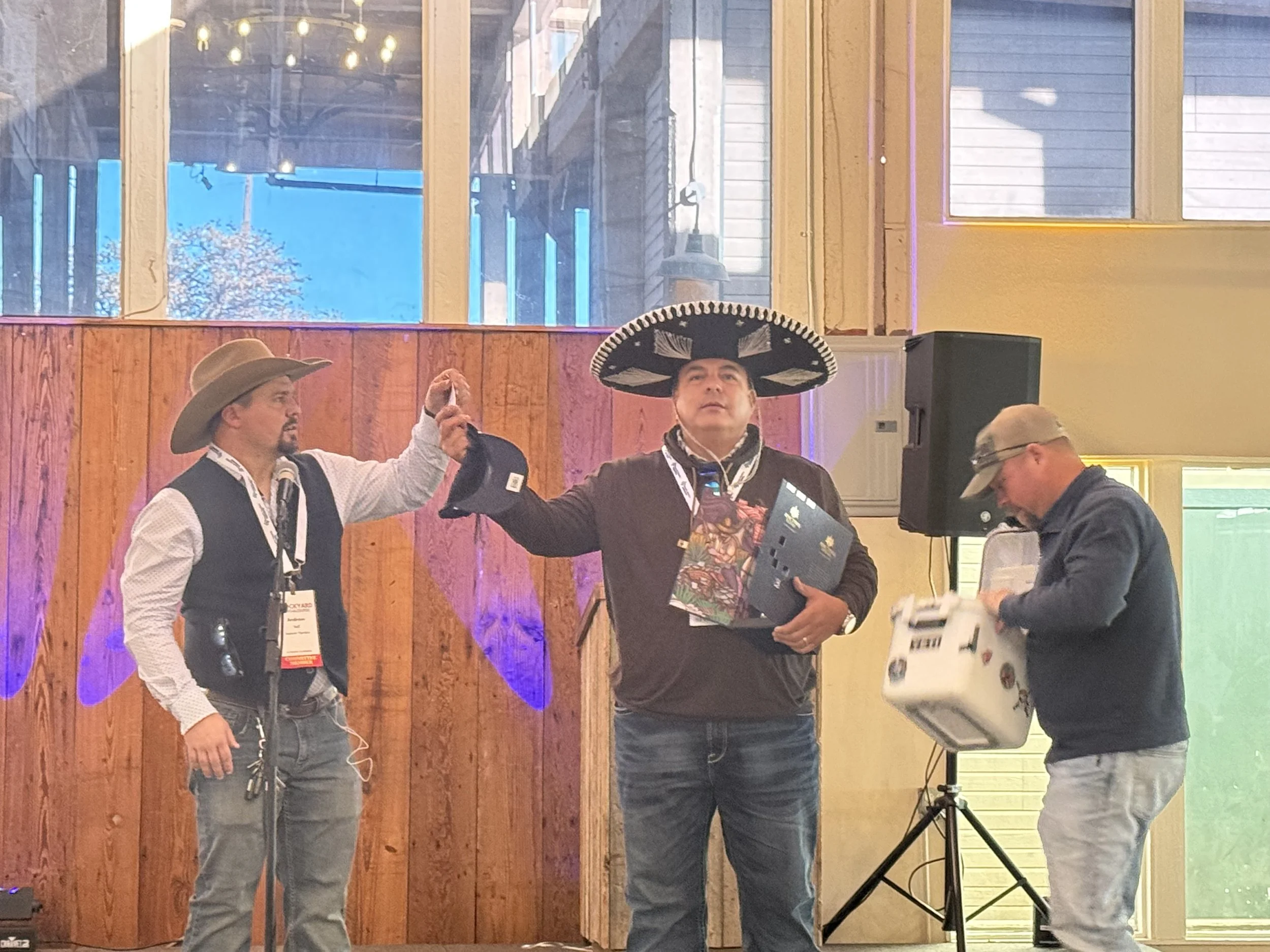 Three men on a stage, two of them wearing large sombrero hats, one dispensing raffle tickets and the other holding various items, with a colorful background and wooden paneling.