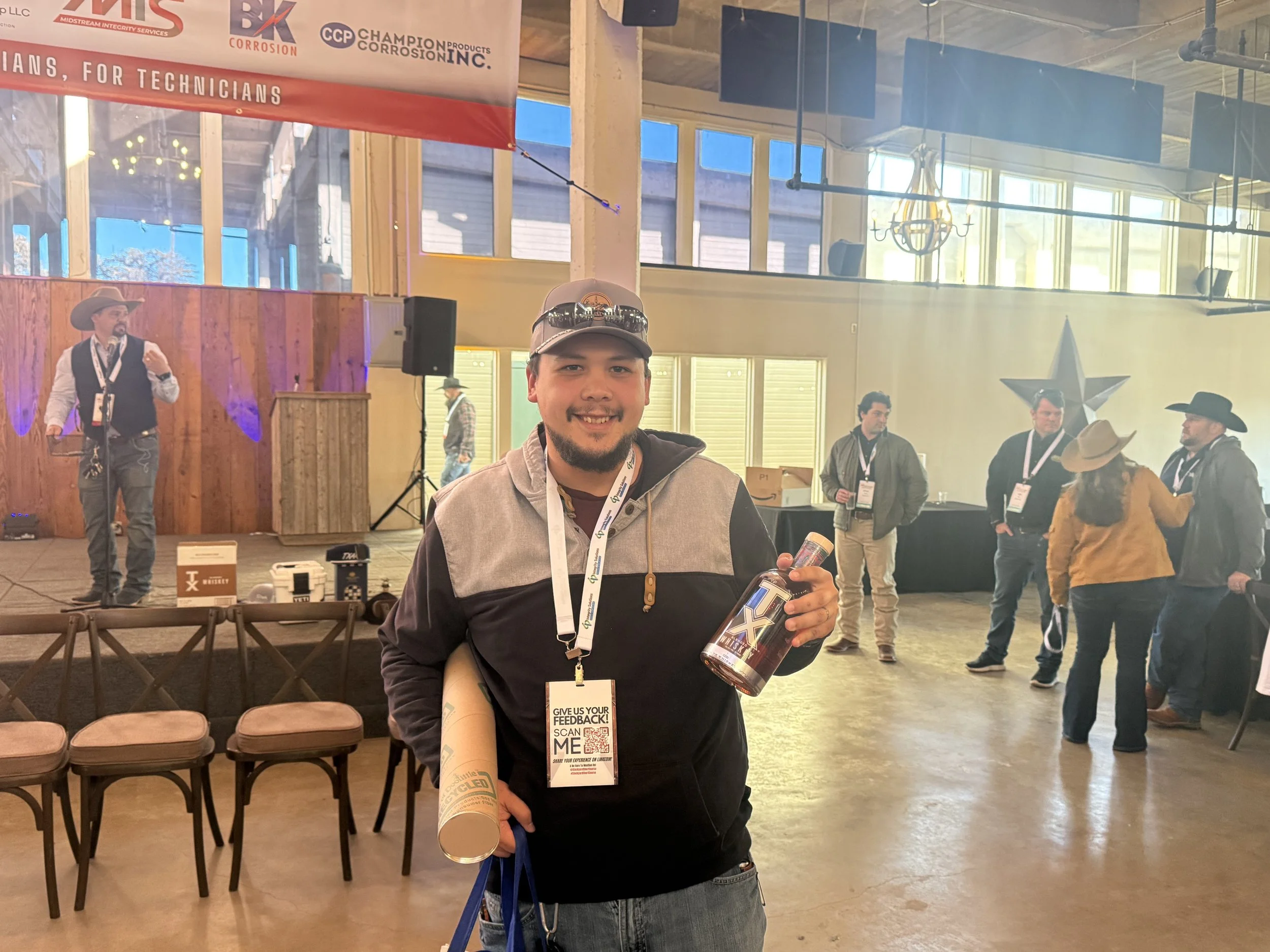 Man smiling holding a T-shirt and a can at an indoor event with a cowboy-themed decor, stage, and other attendees in the background.