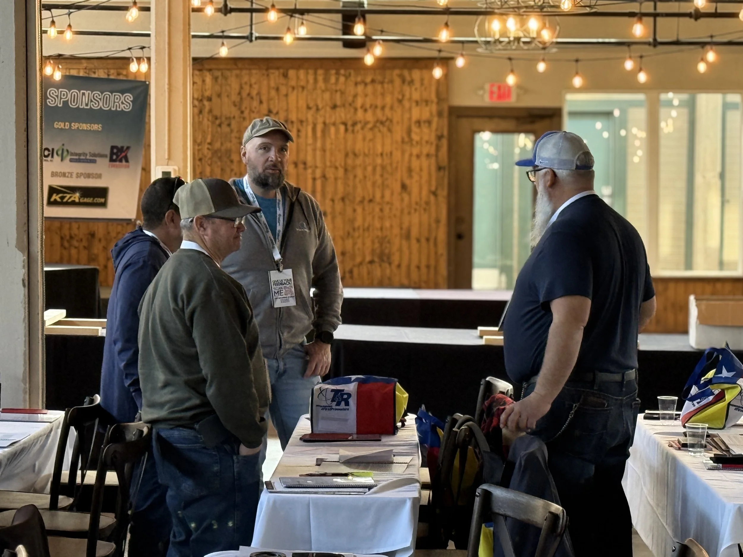 Four men engaged in conversation at a table in a rustic indoor setting with wooden walls and string lights. A sponsor sign is visible on the wall.