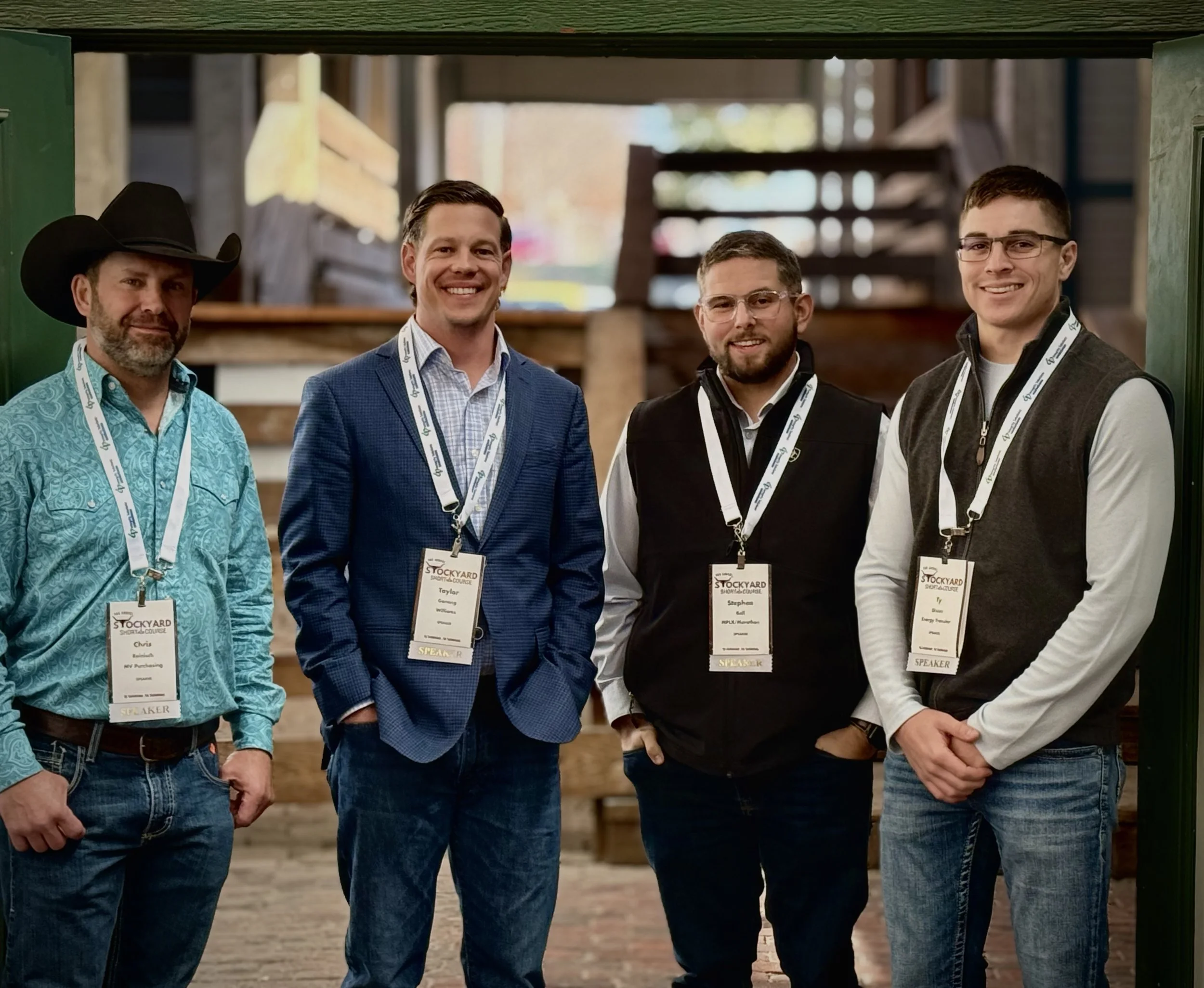 Four men standing indoors, all wearing conference badges and lanyards, smiling at the camera. The background shows wooden structures and a rustic setting.