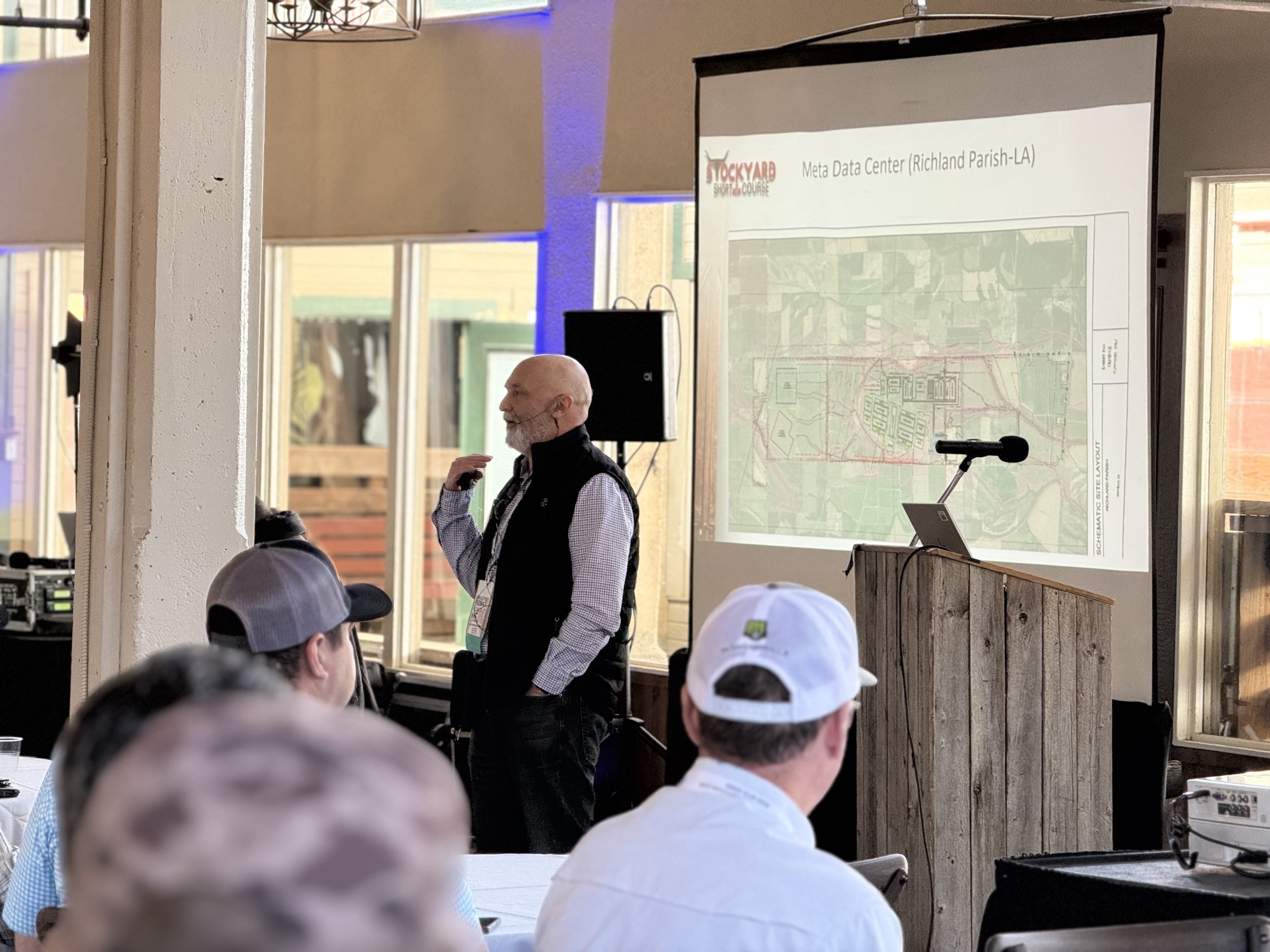 A man with a beard, in a black vest and checkered shirt, is giving a presentation in a room with large windows. A projection screen behind him shows a map titled 'Meta Data Center (Richland Parish-LA)' with the logo 'Stockyard' in the top left corner